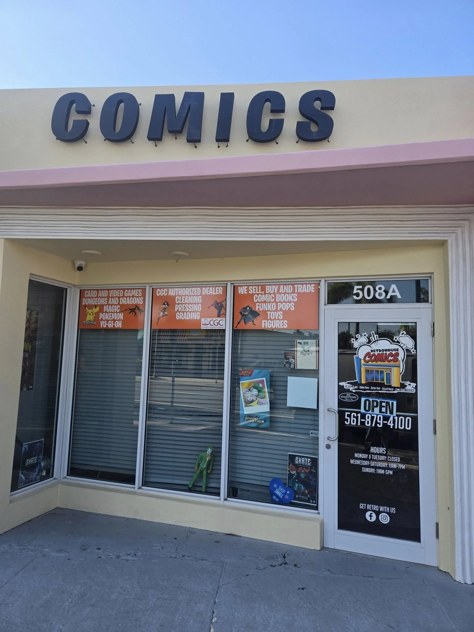 Front of a comic store with a sign that says 'COMICS' and signage indicating they sell comic books, collectibles, and trading cards. Store hours and contact information are visible on the door.