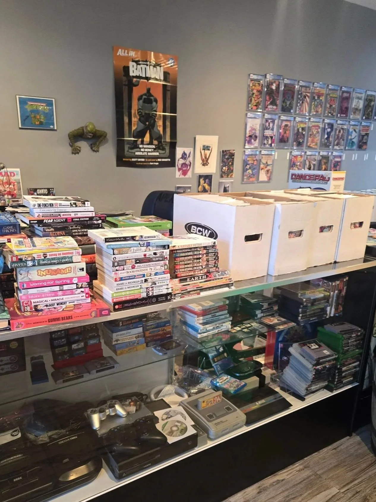 A collection of comic books, video games, and collectibles displayed on a glass counter table and shelves in a room. The room has posters on the wall, including one for Batman and a small figure of a turtle near the wall.