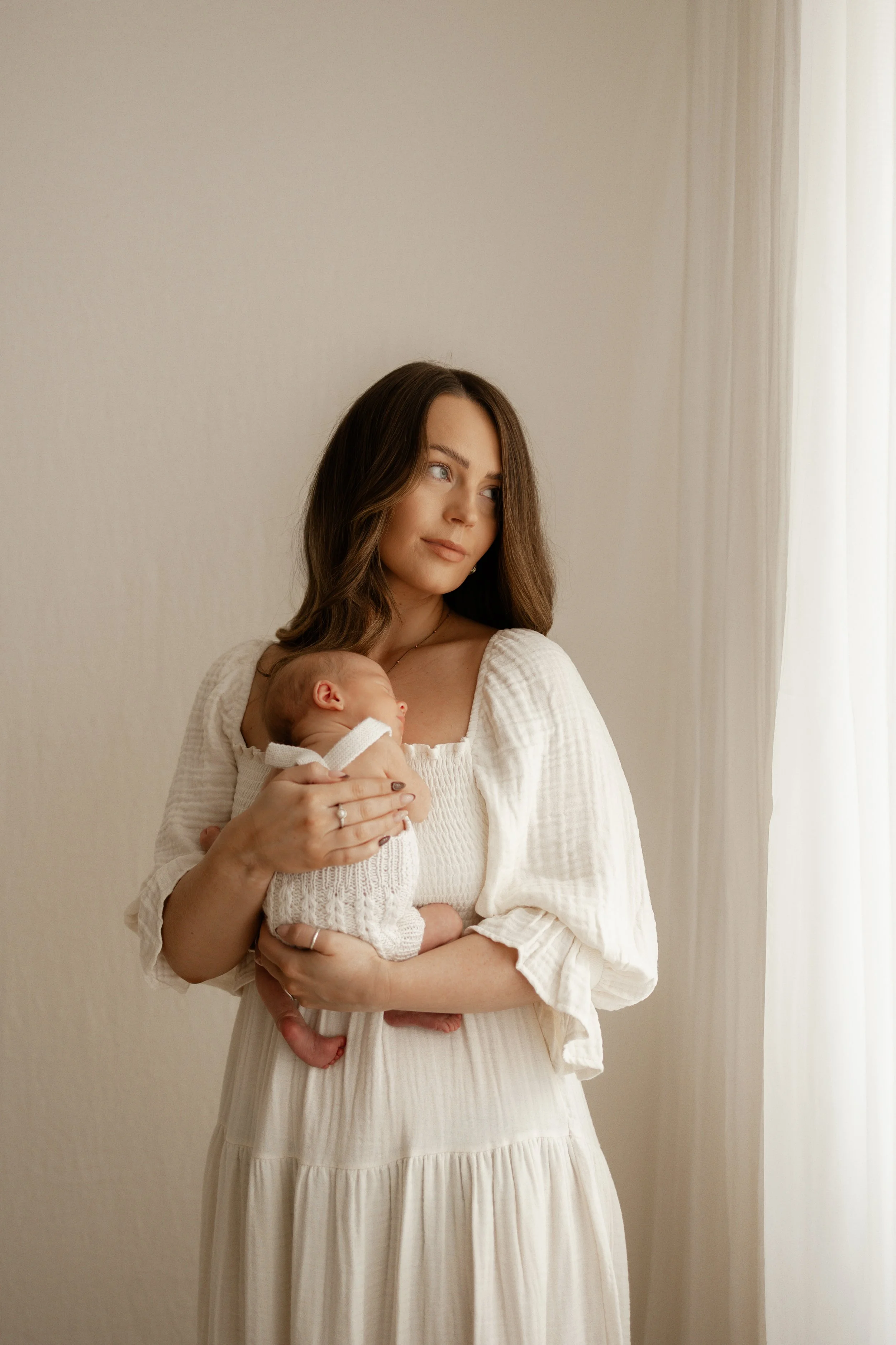 mother glancing out of window filled with beautiful natural light cuddling her newborn baby 