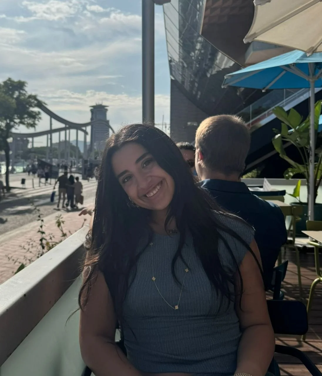 A woman with long dark hair smiling at the camera, sitting at an outdoor cafe with colorful umbrellas, with a city skyline and a bridge in the background.