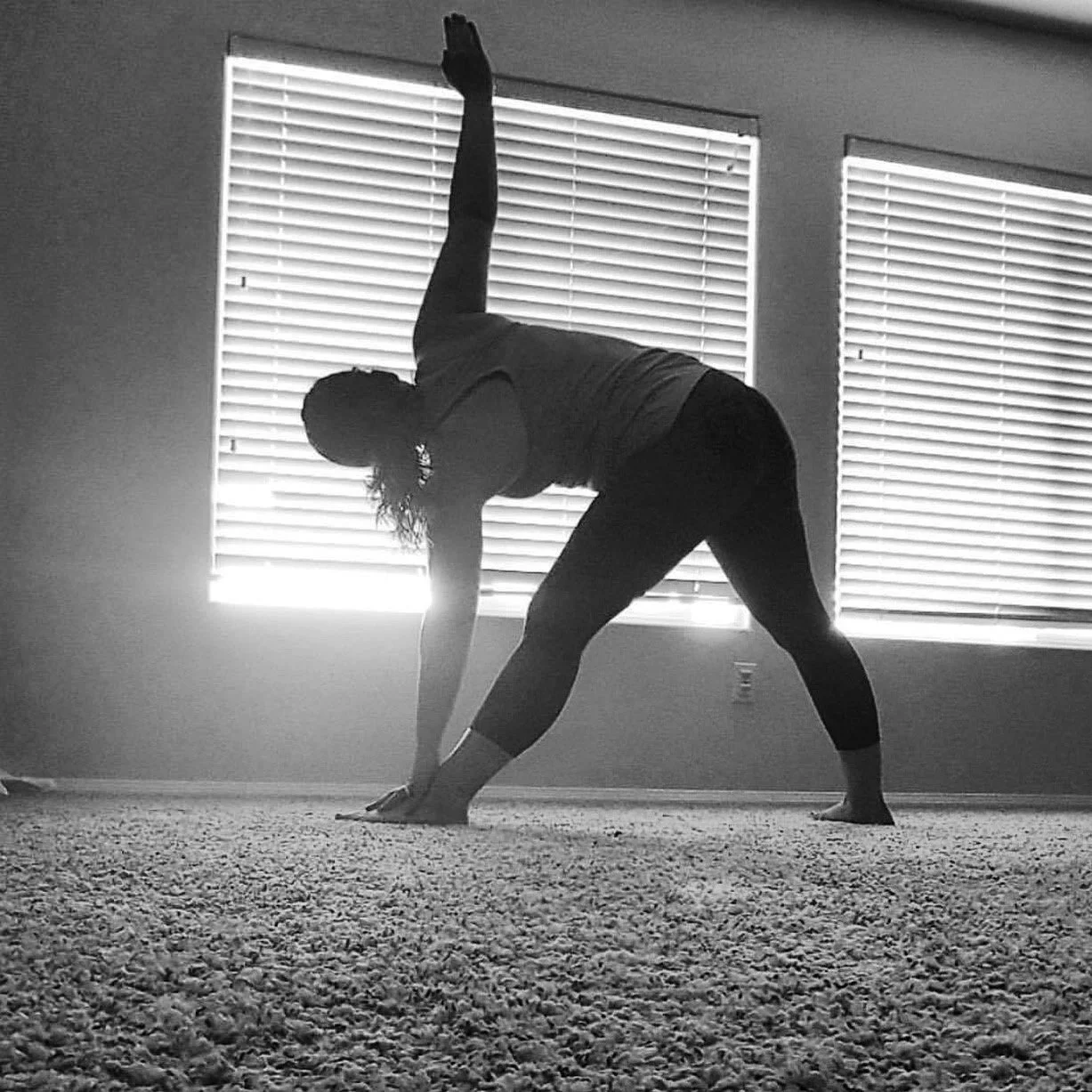 A woman practicing yoga indoors, balancing on her hands and feet with one arm raised upward, in front of two windows with blinds.