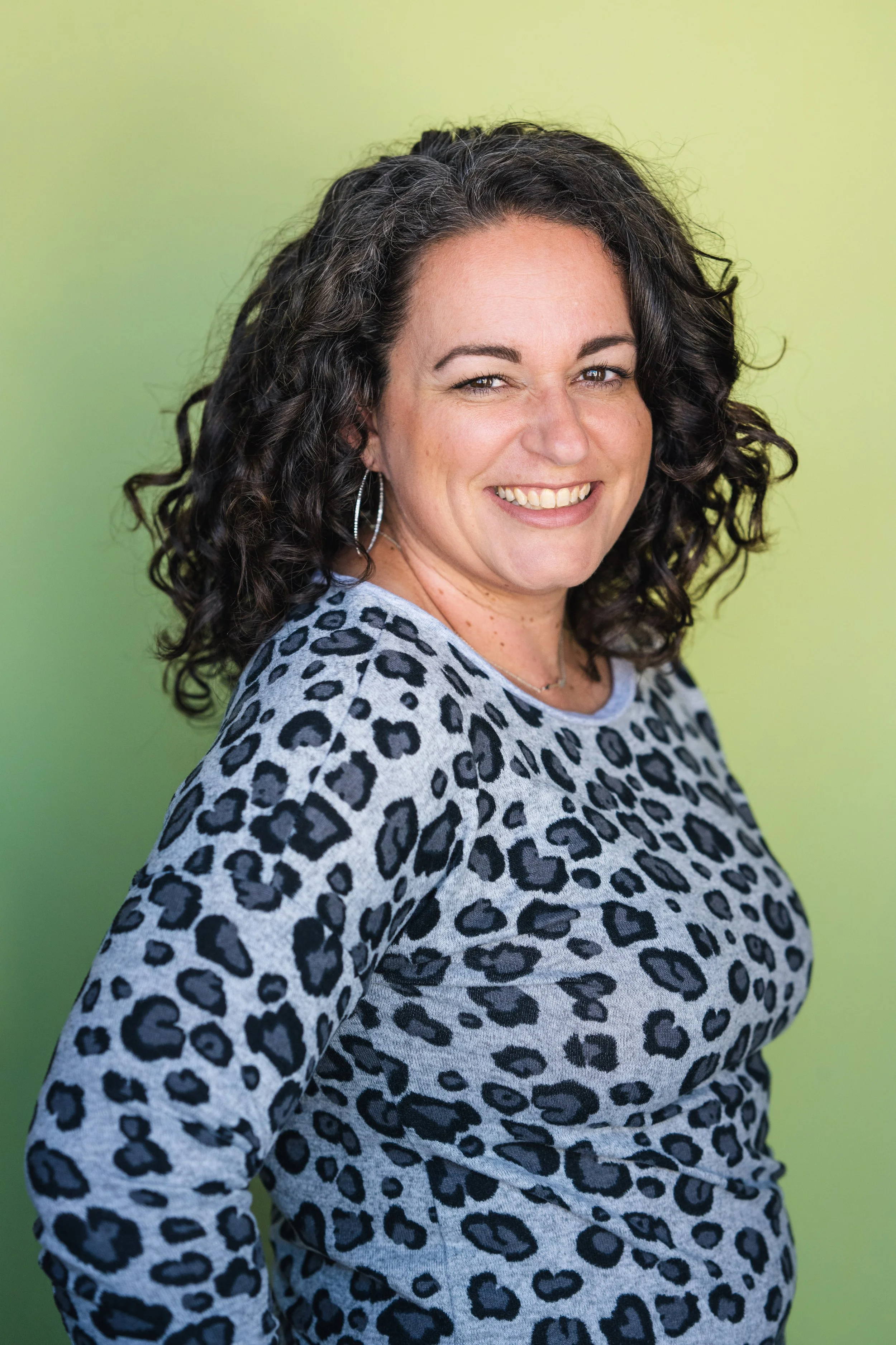 Portrait of a woman with curly dark hair, smiling, wearing hoop earrings and a leopard print top, against a gradient green background.