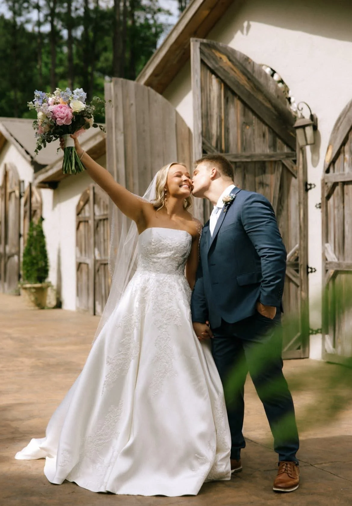 A bride in stunning strapless wedding gown by Veni Infantino with delicate appliques holding her bouquet in the air with excitement while being kissed on the cheek by her husband. Fleur de Vie Bridal Boutique Helena, AL. Birmingham, AL.