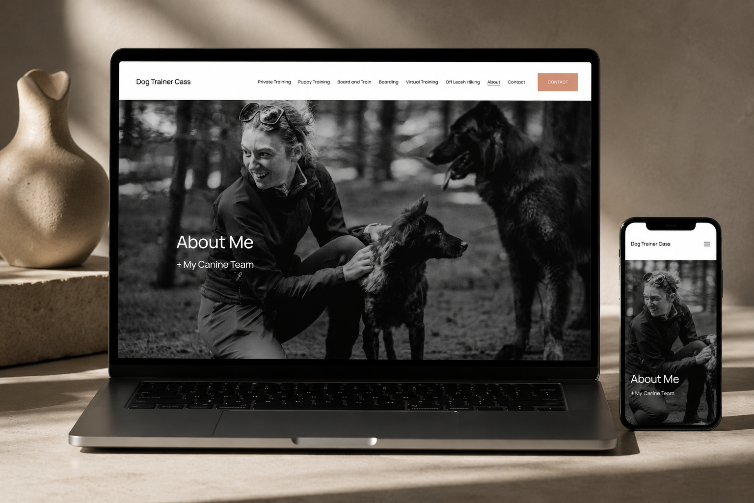 A laptop and smartphone displaying a website about dog training with a black-and-white photo of a woman with glasses kneeling outdoors, interacting with two dogs.