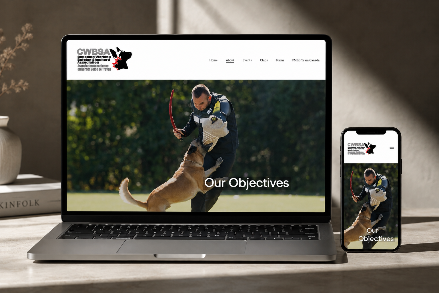 The image shows a laptop and a smartphone on a desk displaying a website titled 'Our Objectives' for the Canadian Working Belgian Shepherd Association (CWBSA). The website features a photo of a person in a protective suit training a Belgian Shepherd dog with a red bite sleeve in an outdoor setting with green foliage.
