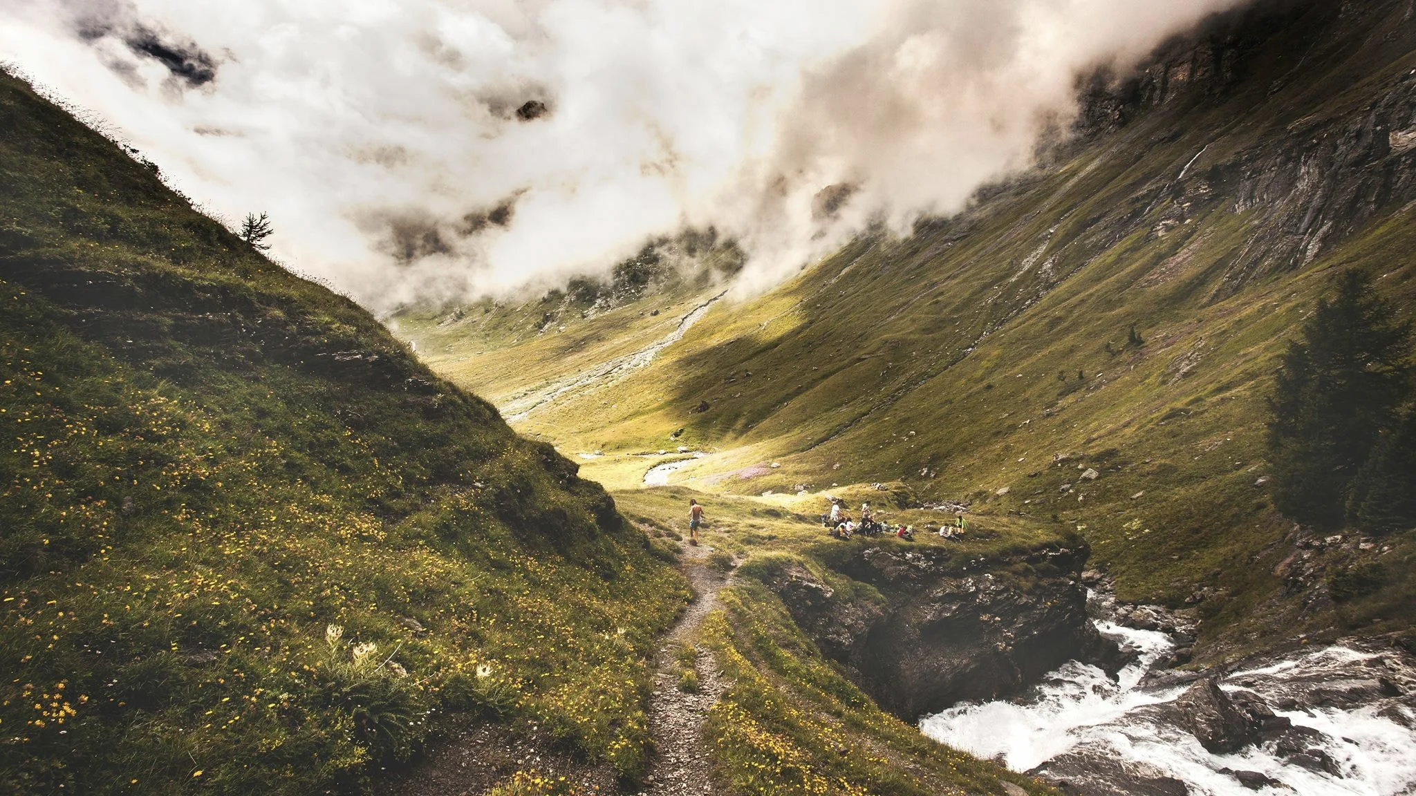 A lush green valley with a narrow stream flowing through, surrounded by tall mountains with clouds above and a group of people resting by the stream.