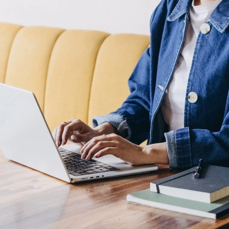Person in a denim jacket typing on a laptop at a wooden table with notebooks and a pen nearby.