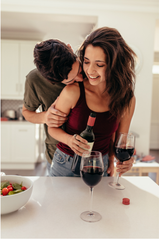 A young woman in a sleeveless red top holding a glass of red wine, with a bottle in her hand, smiling, as a young man behind her kisses her neck in a kitchen.