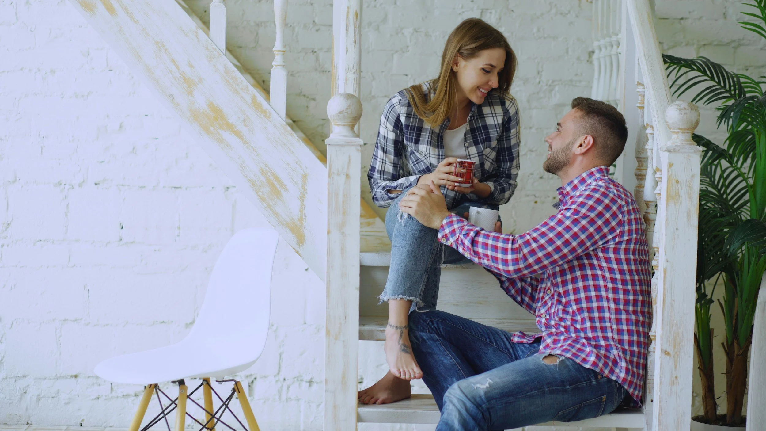 A young man and woman sitting on stairs inside a bright, modern home, sharing a joyful moment. The woman is standing on the stairs, leaning forward, holding a red coffee mug, while the man is sitting on the floor, looking up at her and smiling. There is a white chair with wooden legs to the left and green plants in the background.