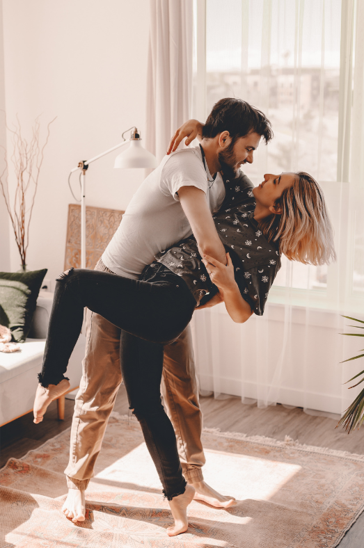 A couple dancing closely in a bright living room, with sunlight coming through large windows.
