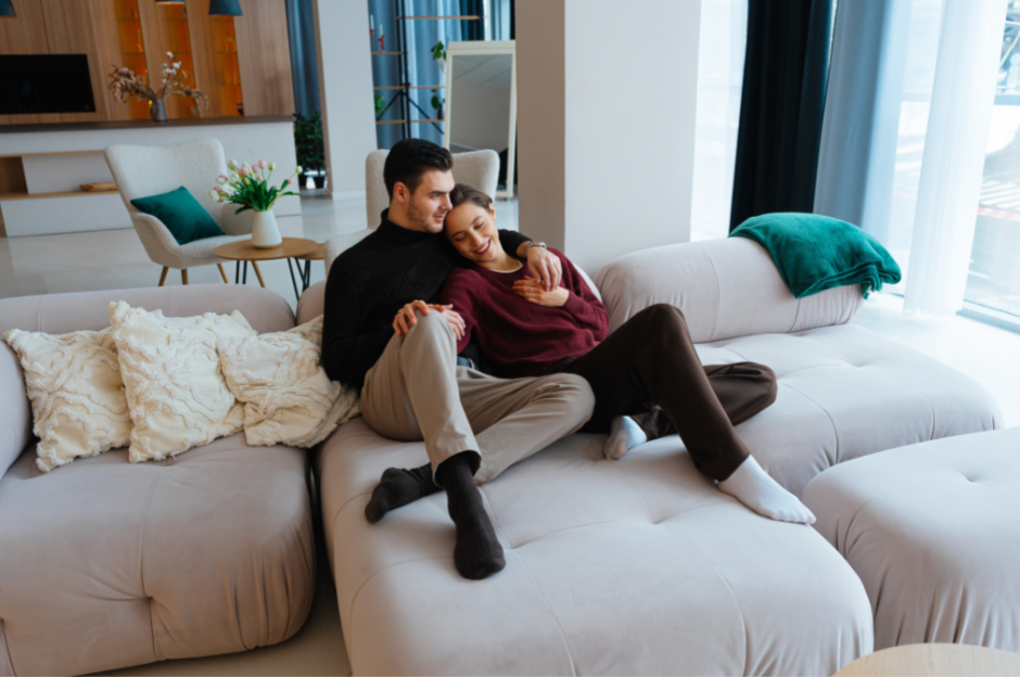 A couple sitting closely on a light-colored sectional sofa in a modern living room, embracing each other and smiling, with curtains and large windows in the background.