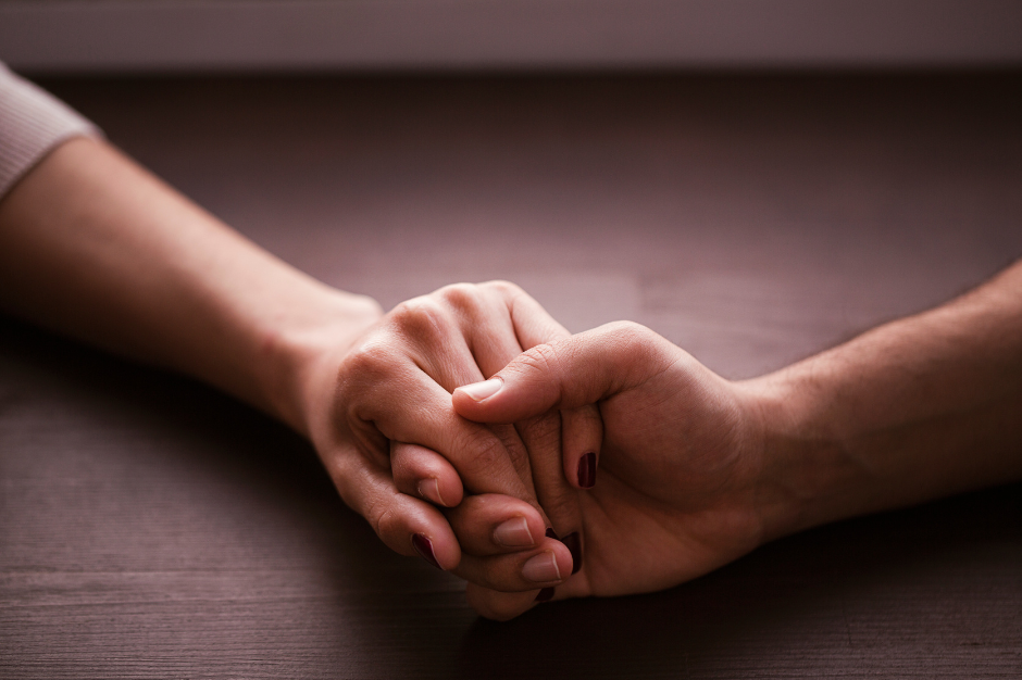 Two people holding hands on a wooden surface, one person with painted nails, engaging in a comforting or supportive gesture.