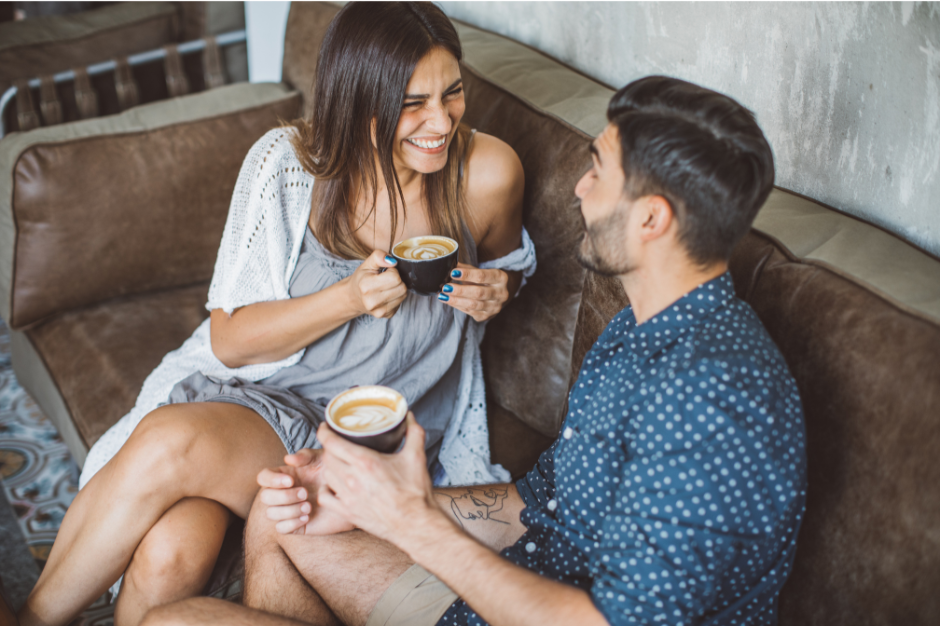 A woman and a man sit on a brown sofa, laughing and enjoying coffee together in a cozy setting.