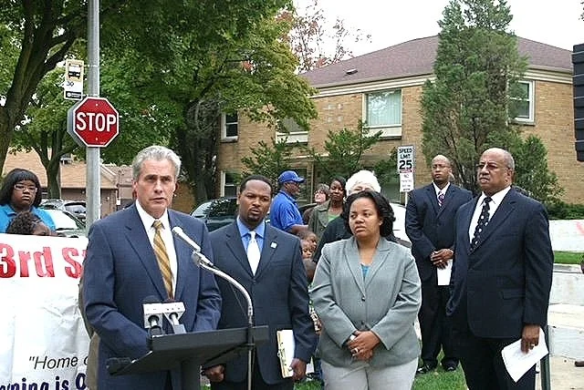 A group of people standing outdoors at a press conference or community event, with a man at a podium speaking. There are trees, a stop sign, and residential buildings in the background.