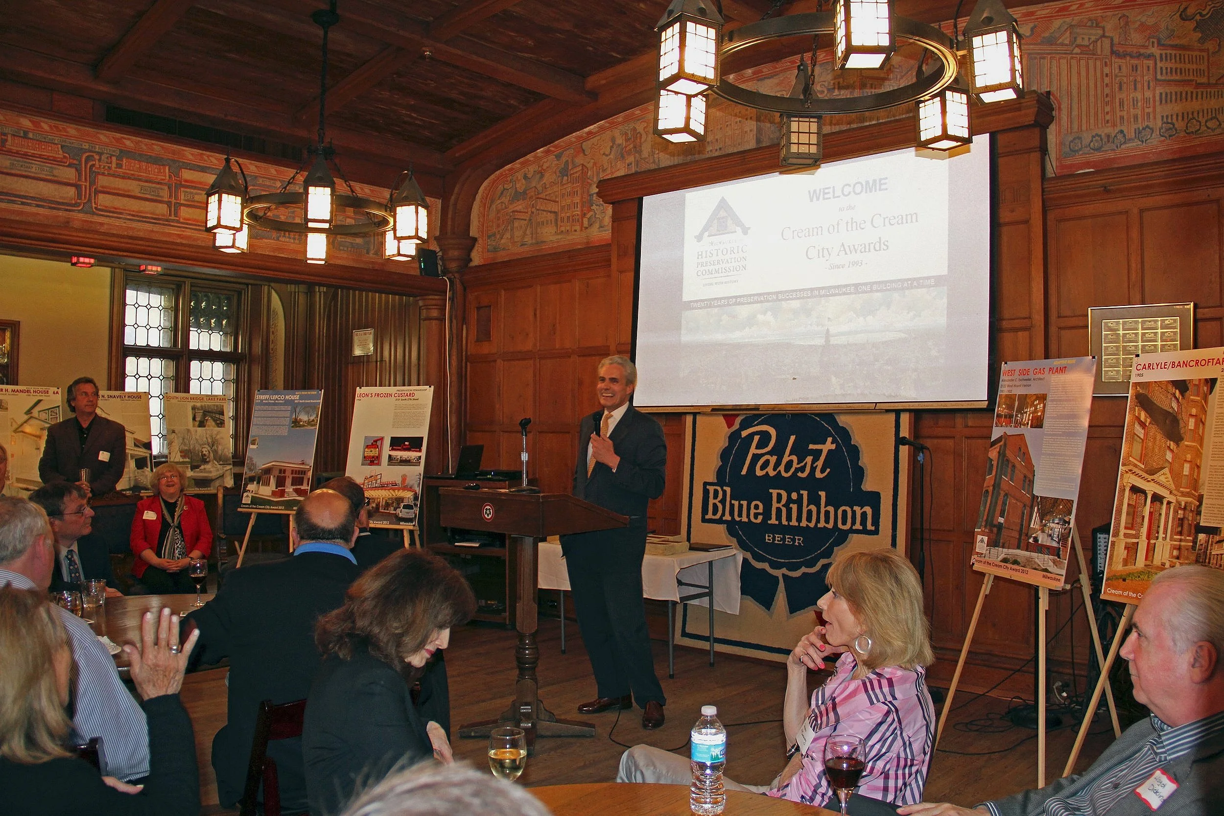 A man in a dark suit standing at a podium, speaking into a microphone at an event inside a wood-paneled room with a large screen behind him. The screen displays a presentation slide titled "Cream of the Cream City Awards". Attendees are seated at tab