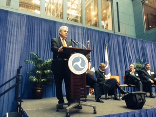 A man at a podium speaking at a conference in front of an audience, with a blue curtain backdrop and sitting panel members.