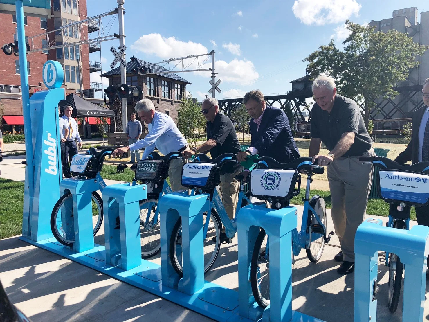 A group of people, mostly men in business attire, participating in a bike-sharing event at a city park. They are standing next to blue bike stations with bikes, with a railroad crossing sign and some buildings in the background under clear skies.