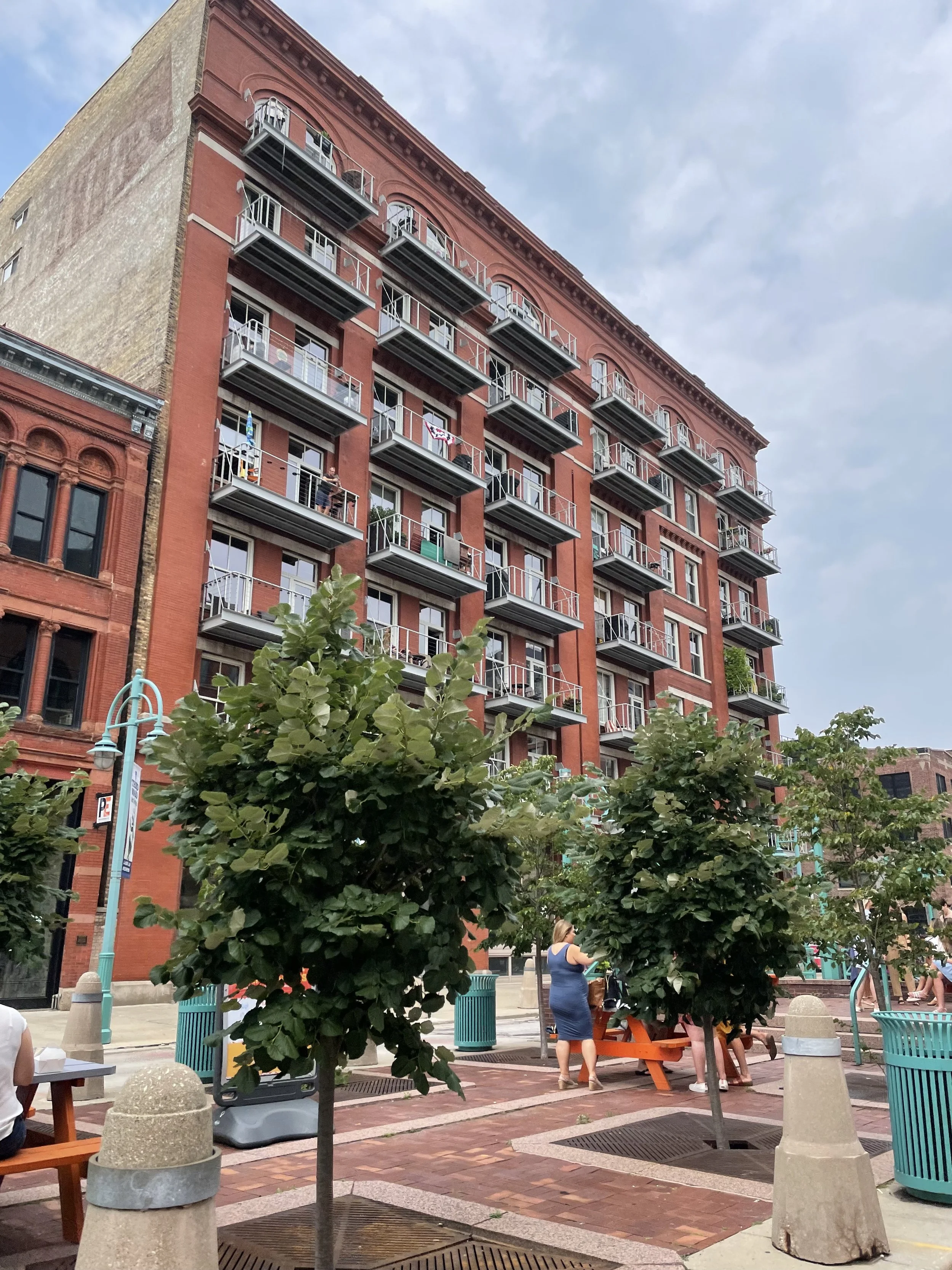 People enjoy a city park with trees, benches, and a brick-paved walkway, with a tall red brick building with balconies in the background under a cloudy sky.