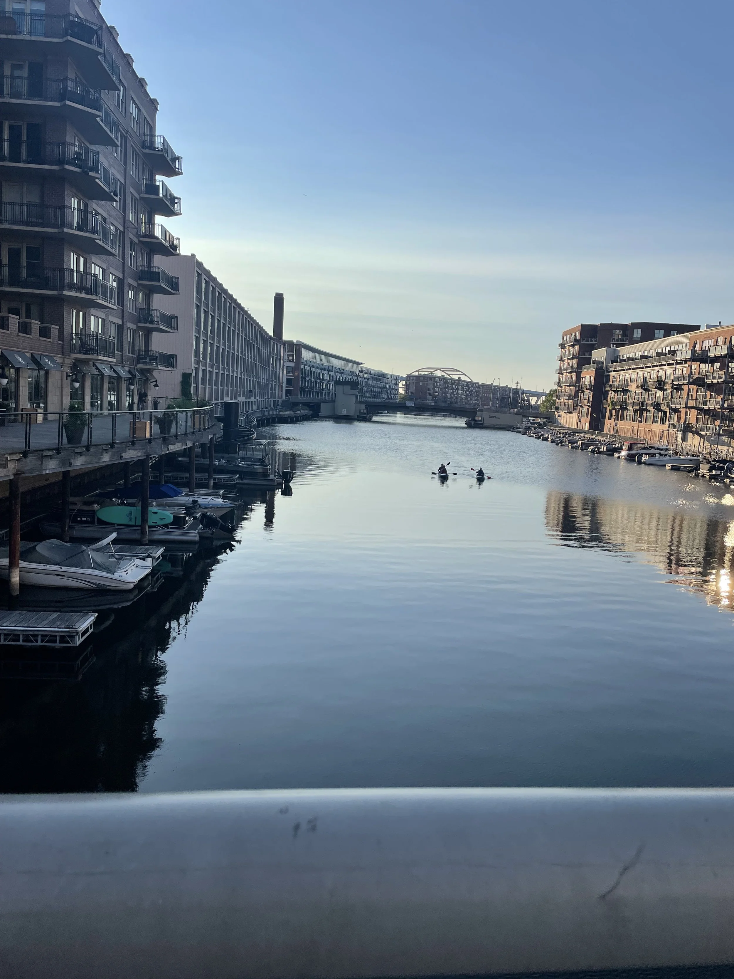 A calm waterway between modern apartment buildings on both sides, with boats docked at the edges and two people kayaking in the distance, under a clear blue sky.