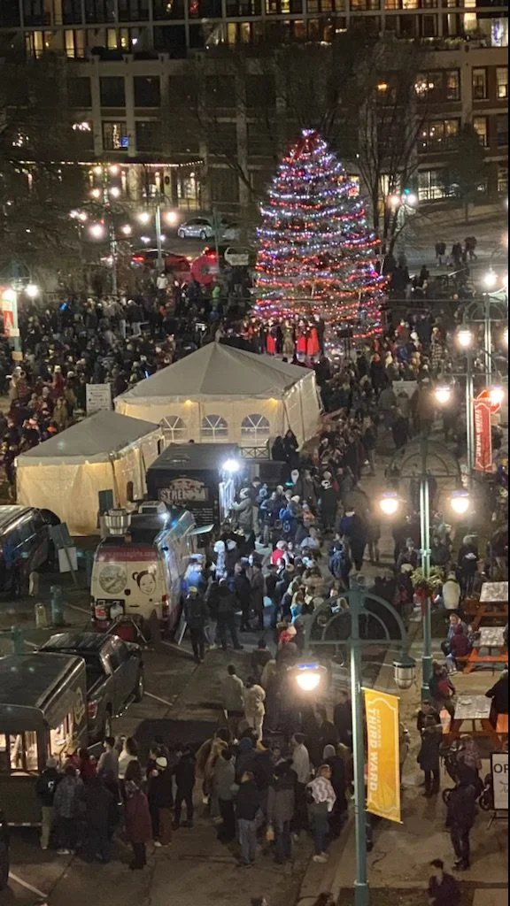 Nighttime scene of a Christmas tree decorated with red, white, and blue lights in a crowded outdoor market. Several tents and food trucks are visible, with people walking and gathered around.