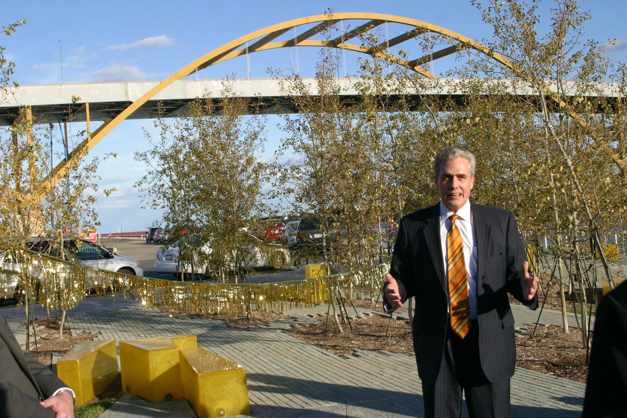 A man in a suit and tie giving a thumbs-up or gesture with both hands, standing outdoors near a decorated area with trees, traffic and a large yellow arch bridge in the background.