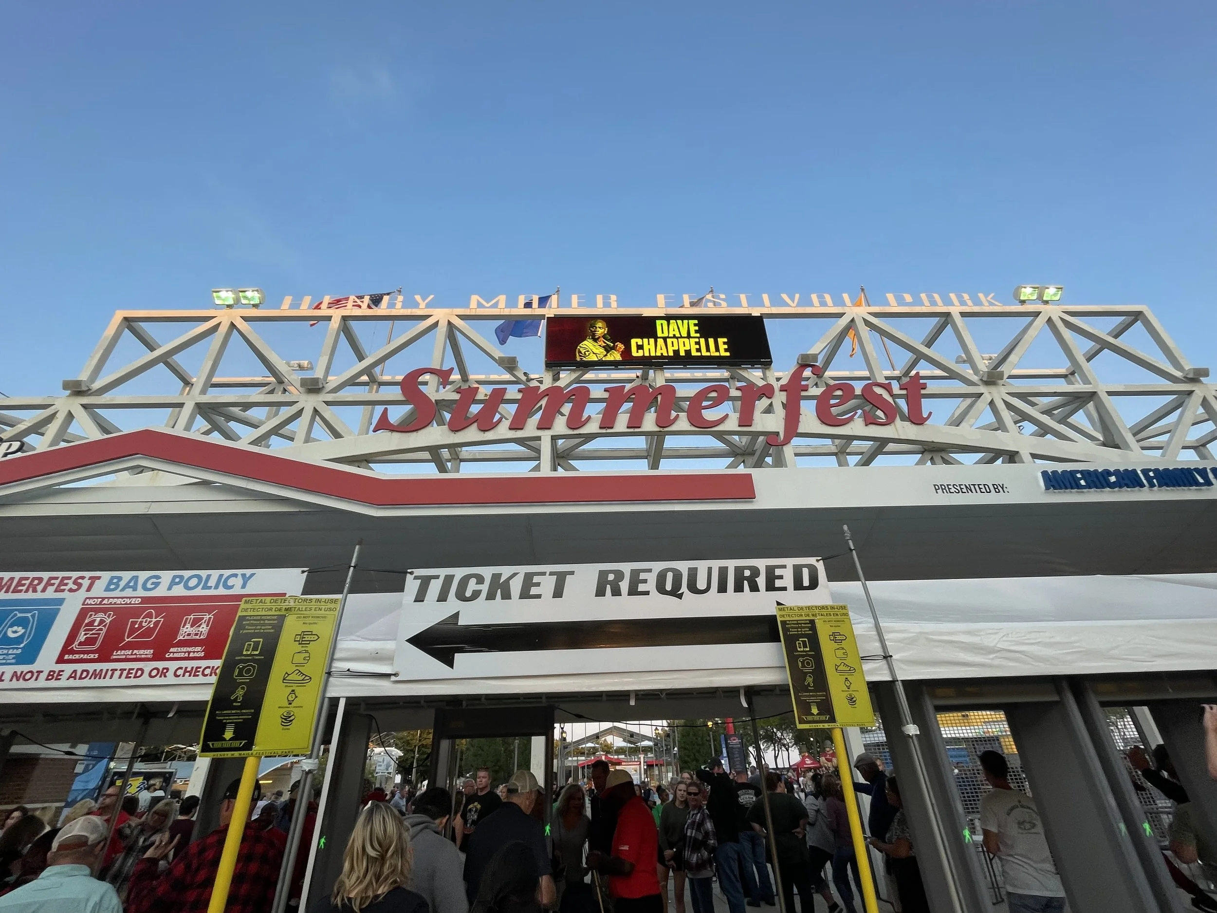 Entrance to Summerfest with a crowd of people, signs indicating ticket requirement and bag policy, and a display board featuring Dave Chappelle.