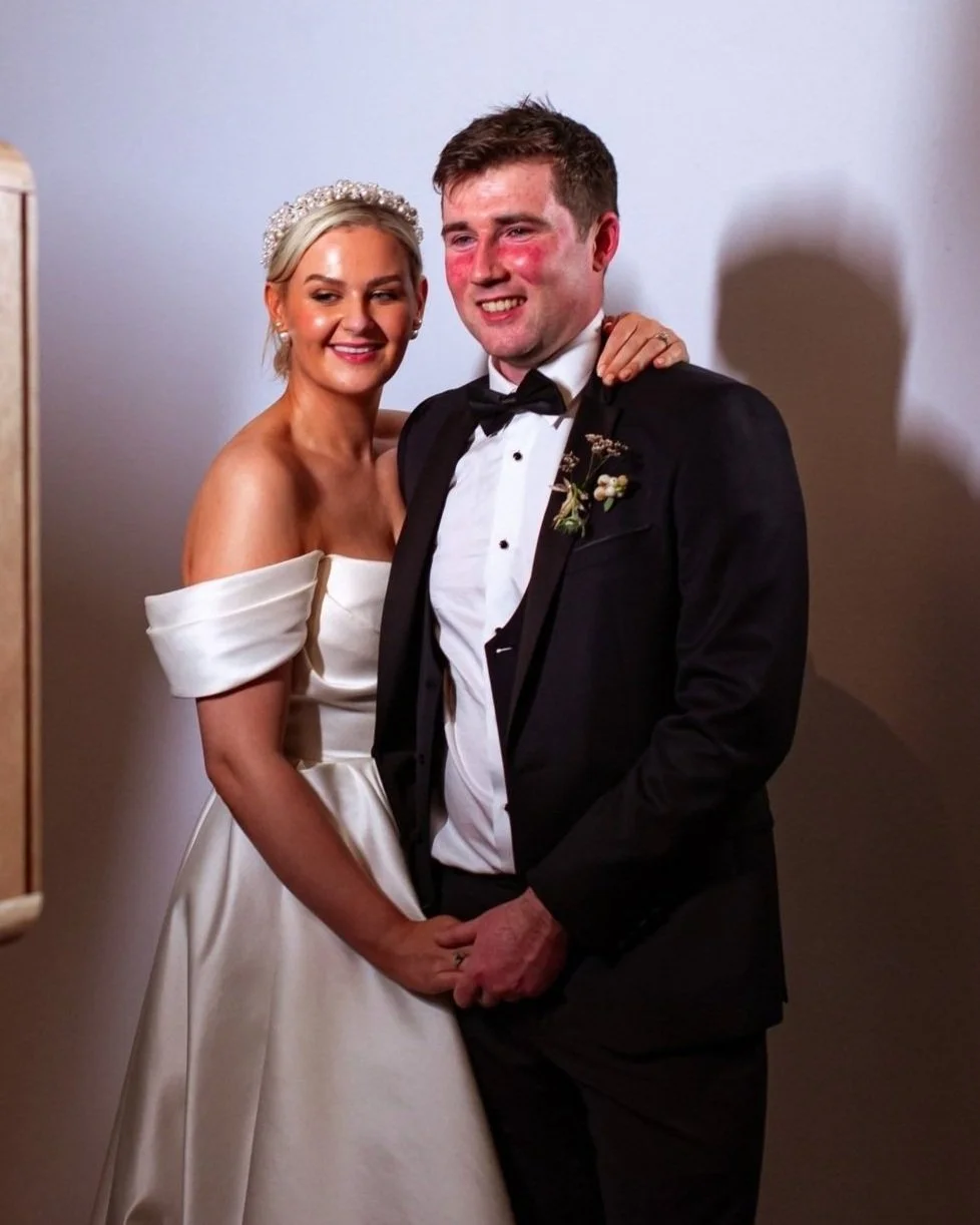 A bride and groom pose for a wedding photo indoors. The bride is wearing an off-the-shoulder white satin wedding gown with a pearl headband. The groom is dressed in a black tuxedo with a bow tie, and has a boutonniere on his left lapel. They are smiling and holding hands, standing close together.
