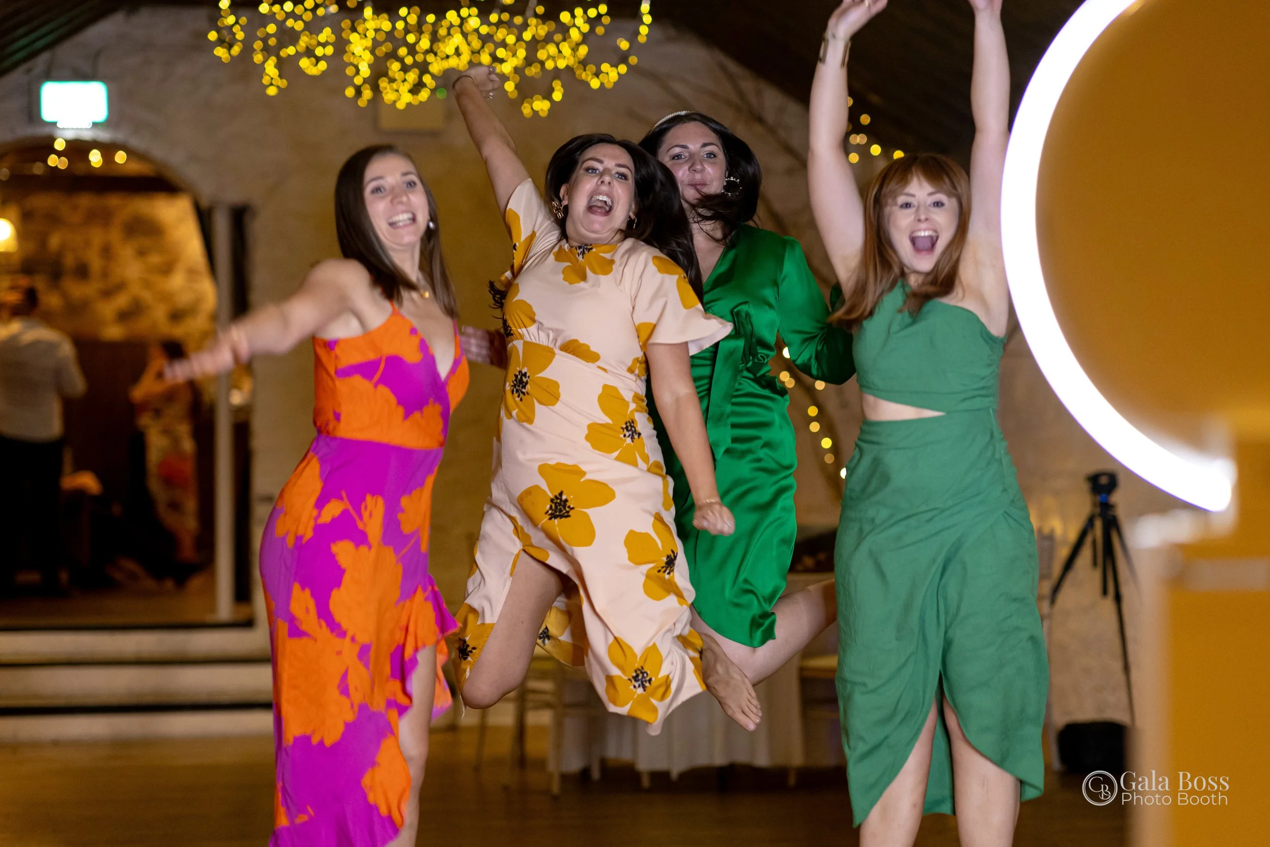 Four women in colorful dresses joyfully jumping and celebrating indoors, with fairy lights hanging from the ceiling and a camera tripod in the background.