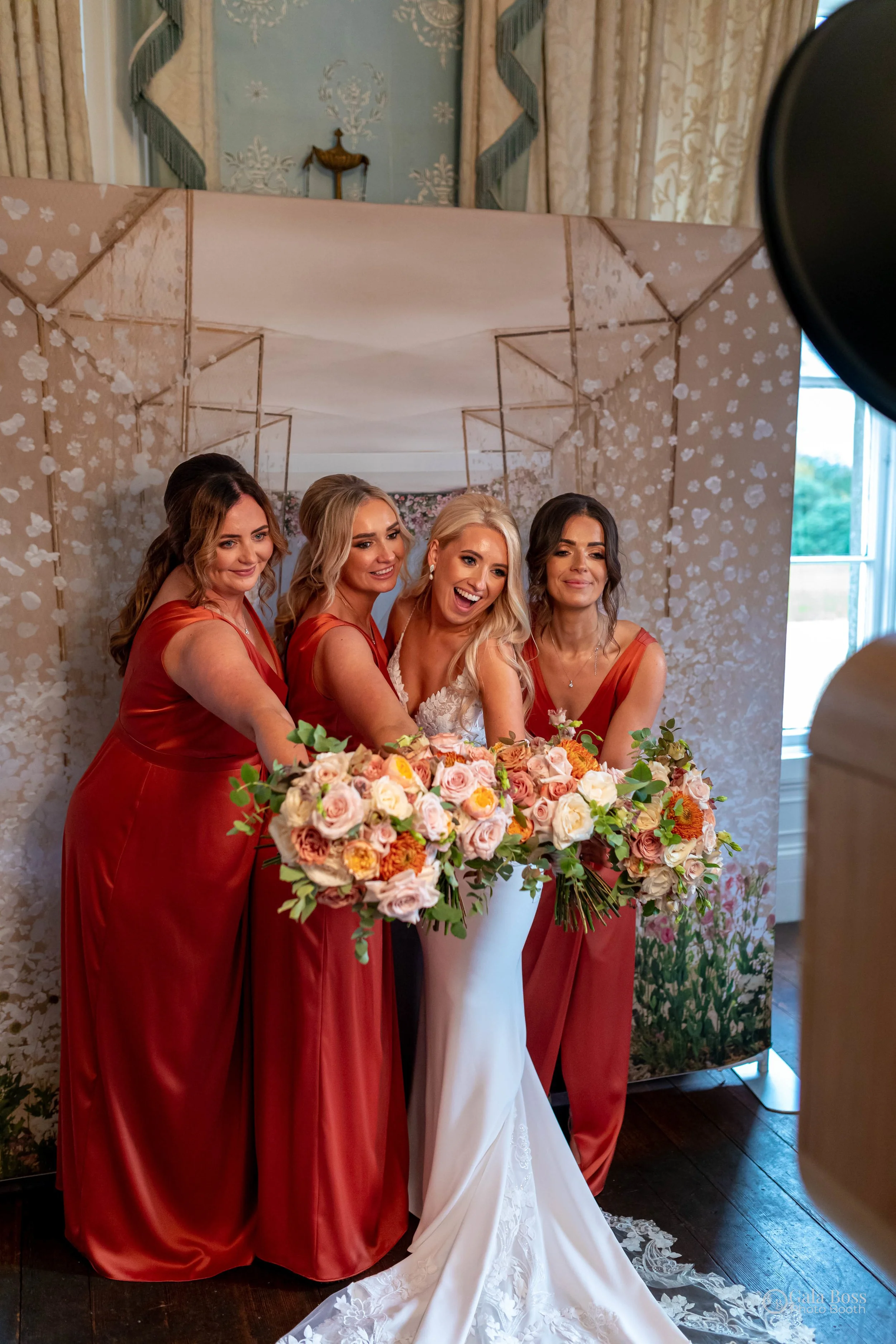 A bride in a white wedding dress and four women in matching red dresses holding large flower bouquets, standing in front of a decorative backdrop during a wedding event.