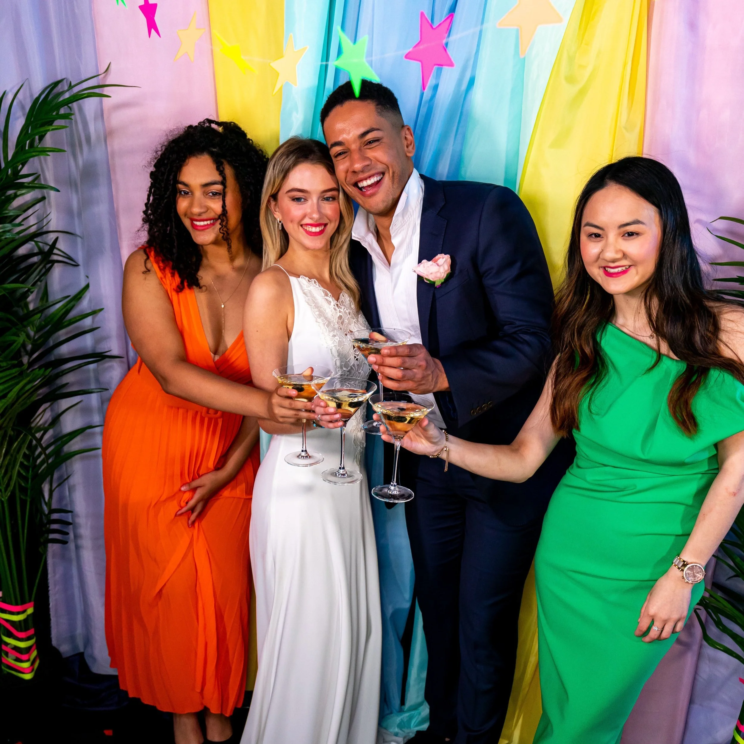 Four people dressed in formal attire, holding martini glasses, standing in front of colorful, star-decorated backdrop.