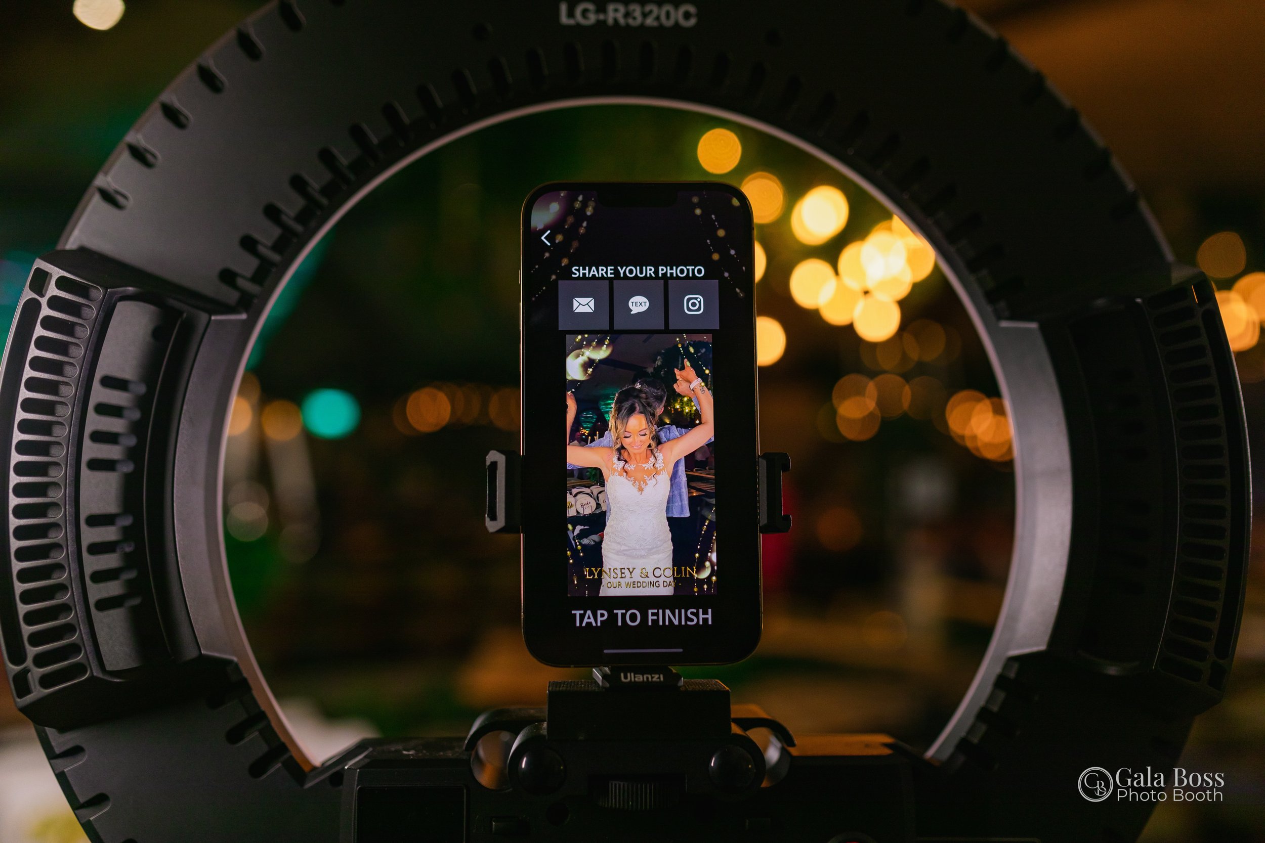 A smartphone mounted on a light ring captures a photo of a bride at a nighttime wedding celebration, with blurred string lights in the background.