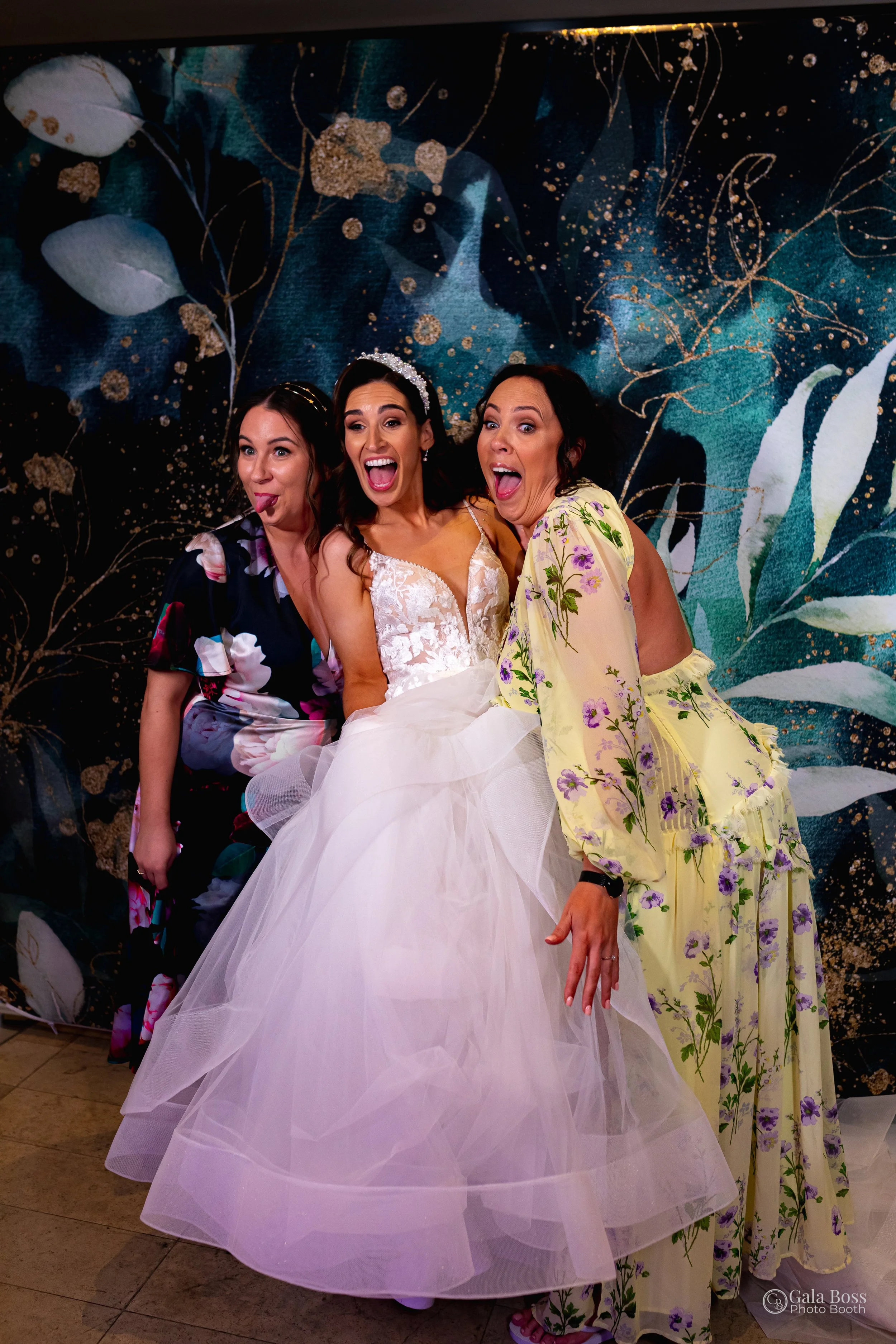 Three women posing playfully at a wedding, with one in a bridal gown and two in floral dresses, standing in front of a decorative backdrop.