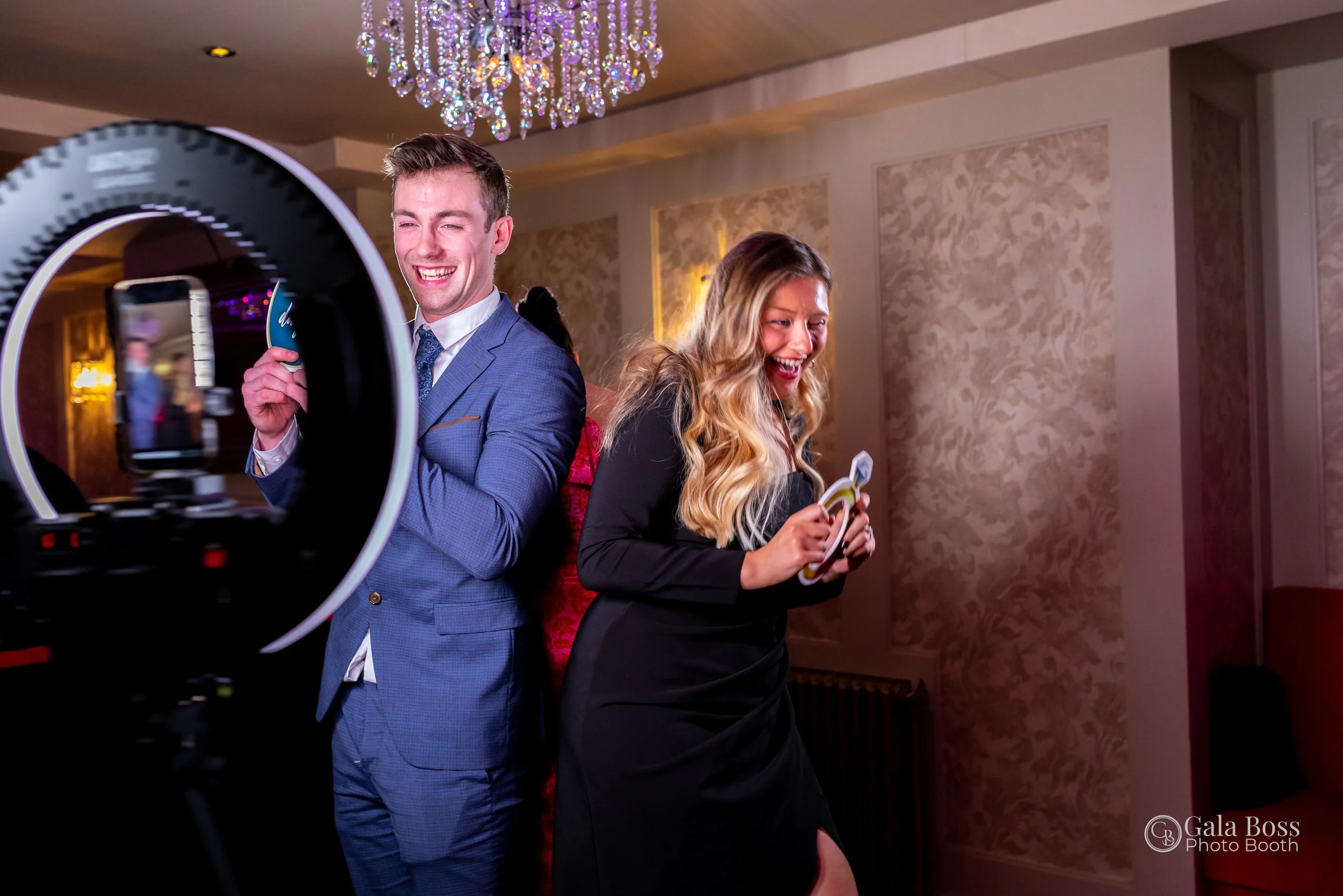 Two people laughing and holding props in front of a ring light at a photo booth, with a chandelier overhead and decorated walls.
