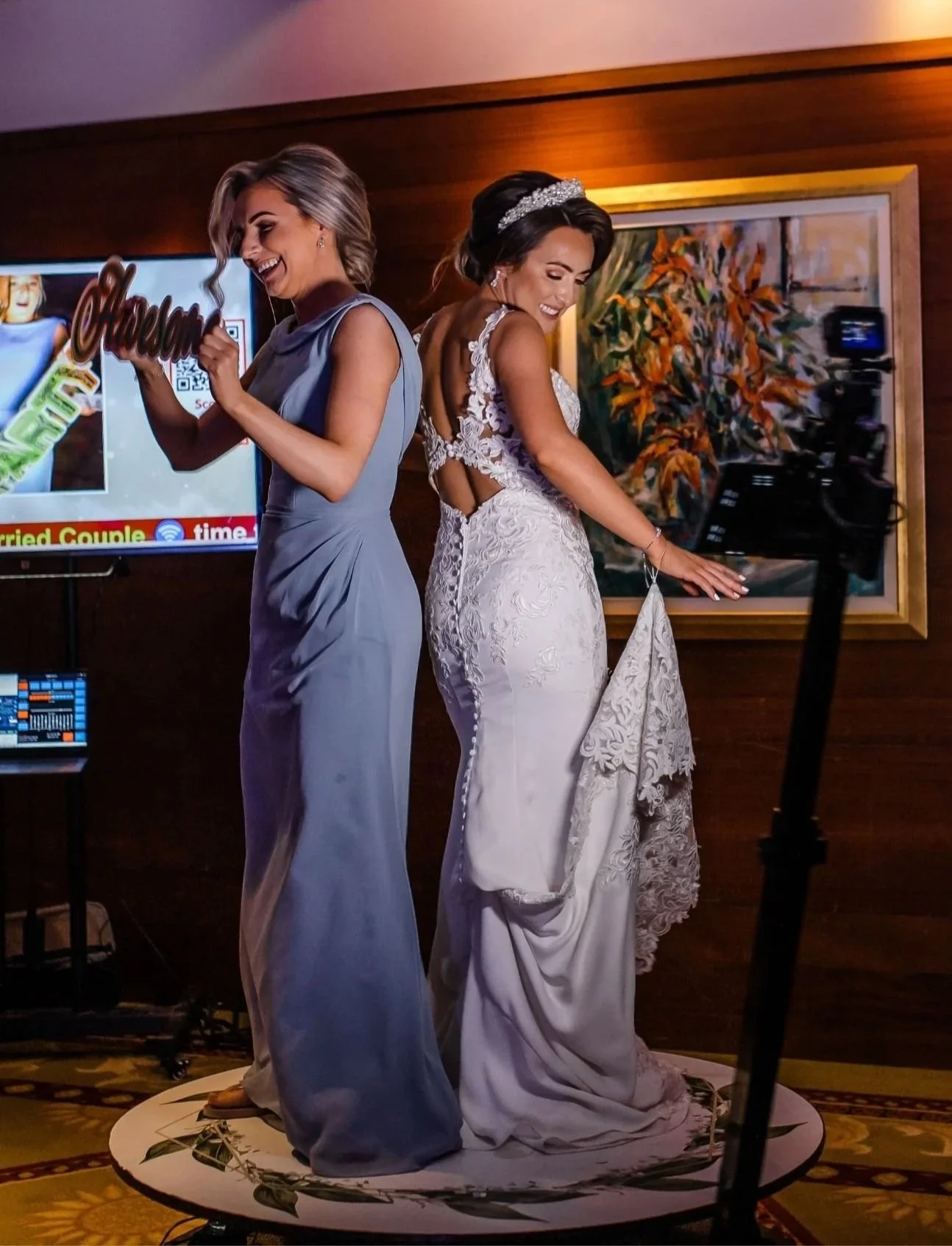 Two women in elegant dresses smiling and dancing together at an indoor celebration, with a television screen and artwork in the background.