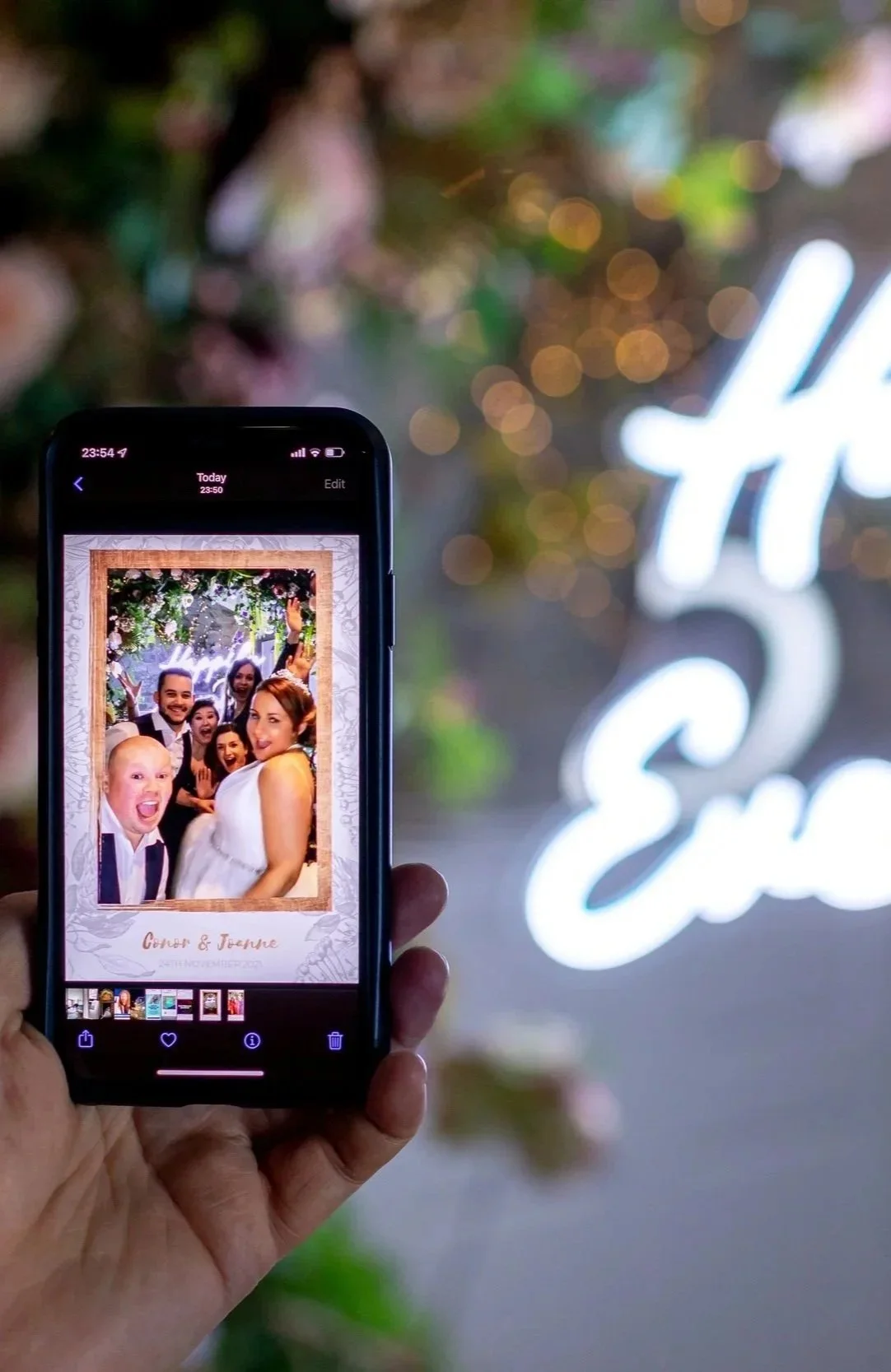 A person holding a smartphone displaying a group photo of wedding guests in front of a floral backdrop, with blurred decorative lights and a neon sign in the background.