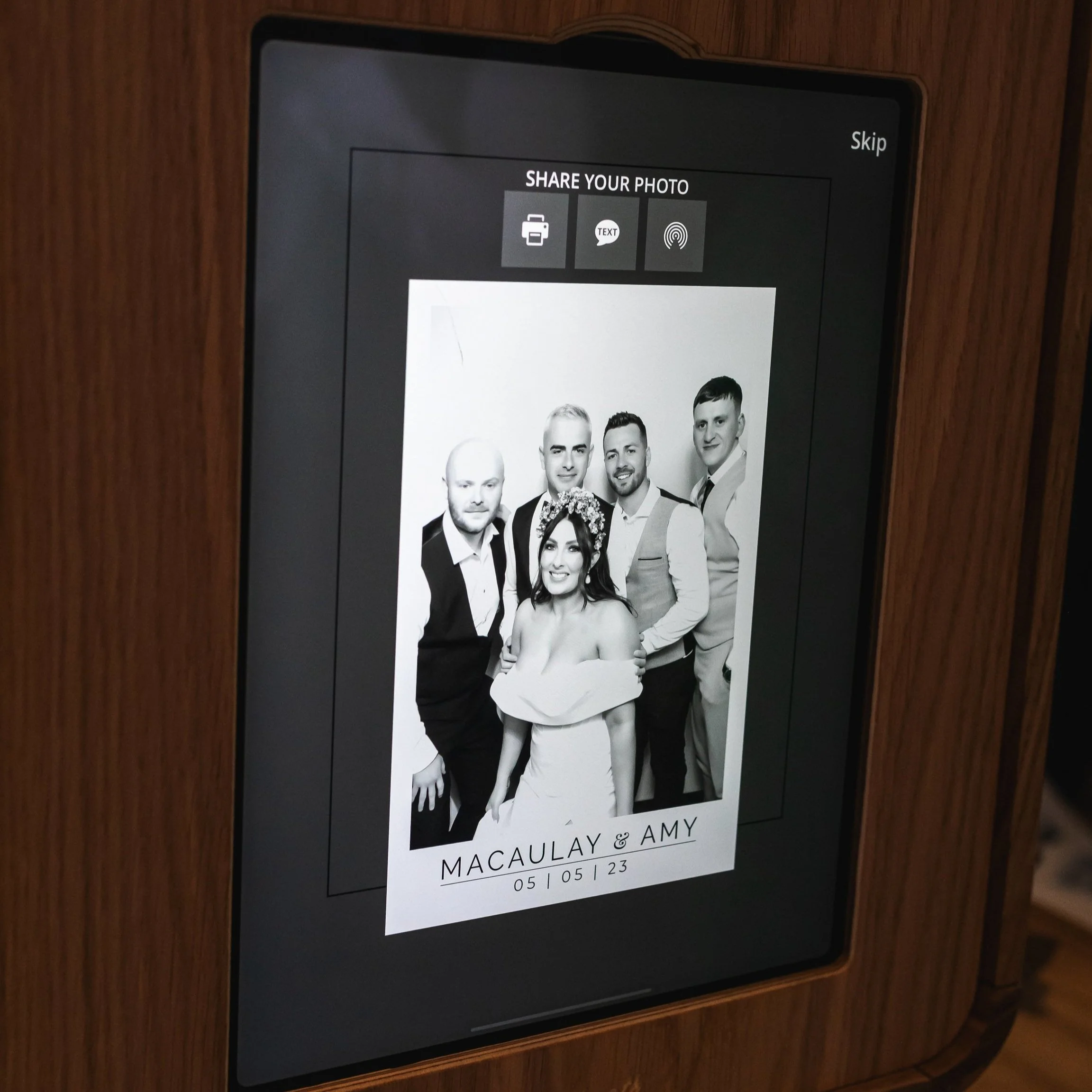 Photo booth screen displaying a black and white photo of five people, four men and one woman, at a wedding event. The woman is wearing a strapless dress and a floral crown. Text at the bottom reads "MACAULAY & AMY  05 | 05 | 23".