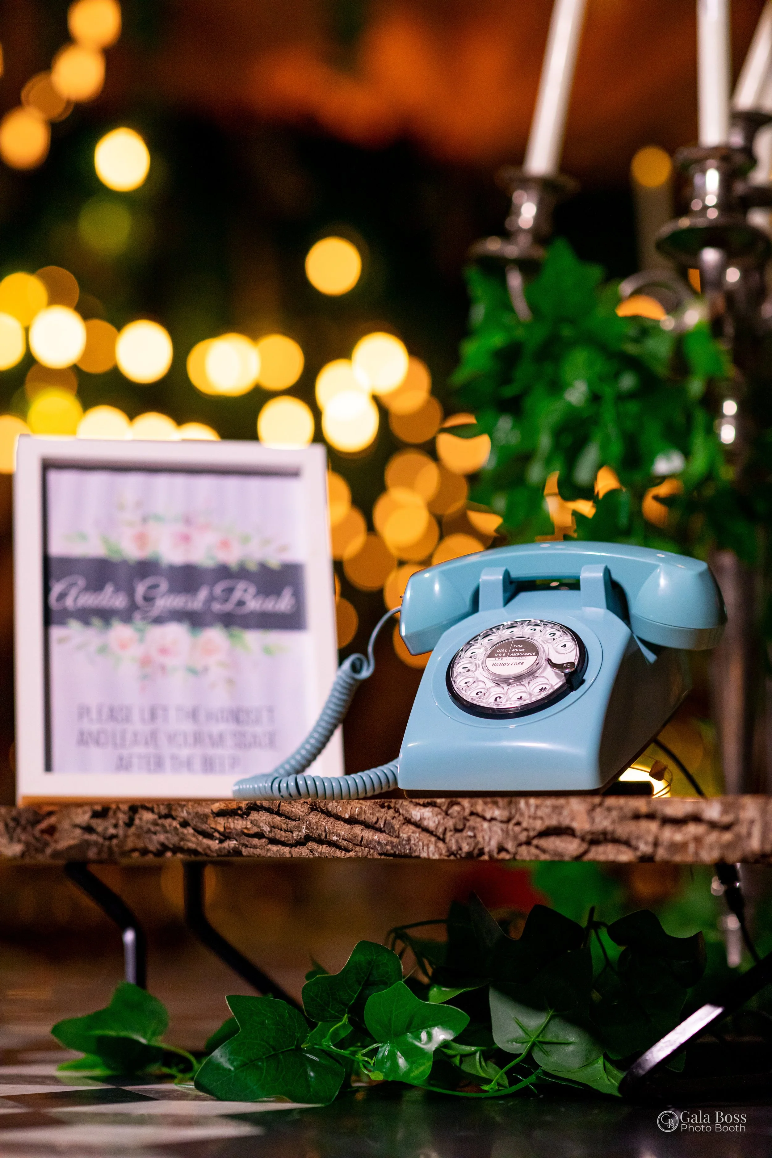 A vintage blue rotary phone on a wooden table with a blurred floral arrangement and string lights in the background.