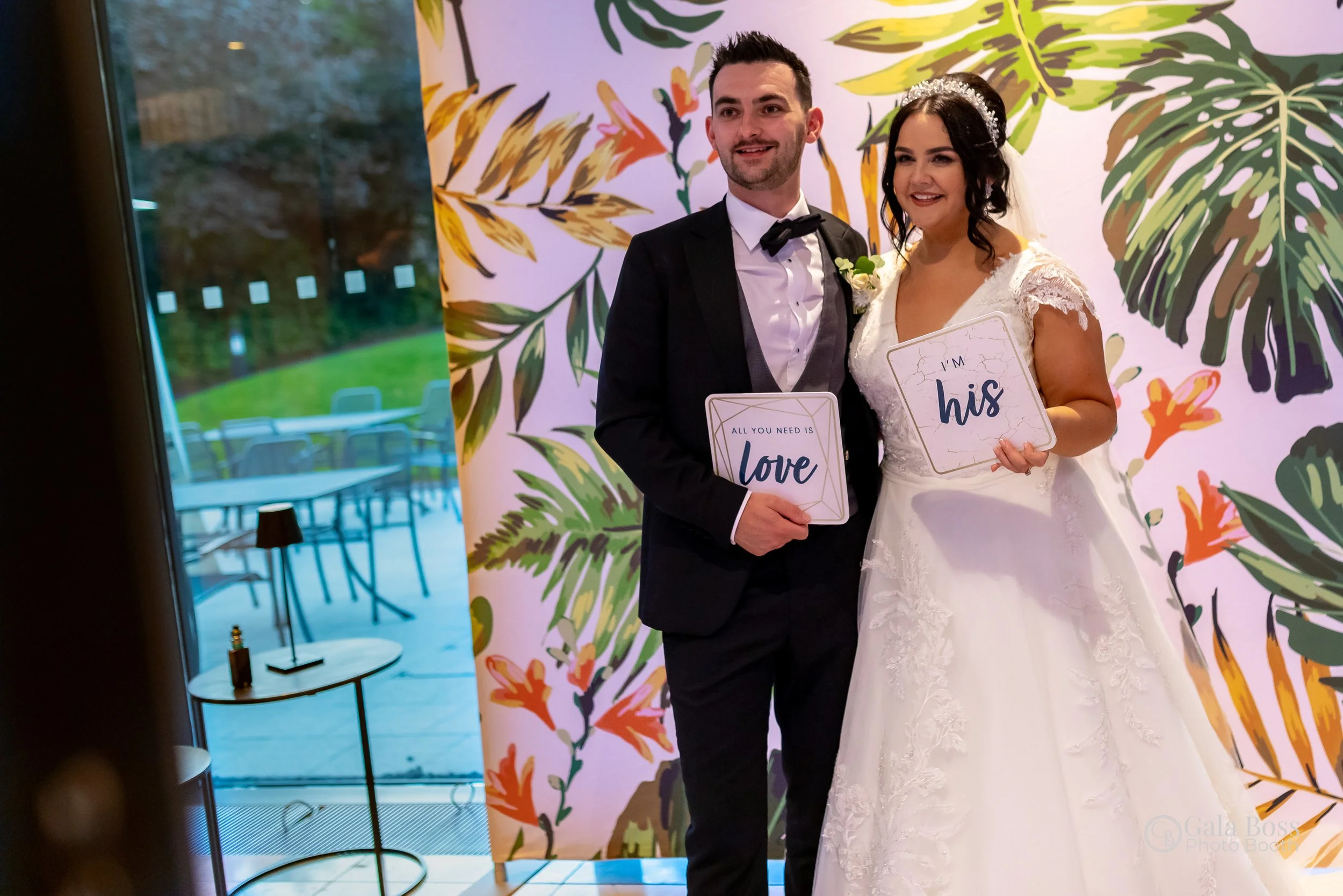 Bride and groom holding signs reading "All you need is love" and "I'm his" standing in front of a colorful floral backdrop at a wedding.
