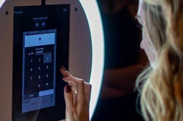 A woman using a touchscreen photo booth with a ring light, entering a phone number on the screen.
