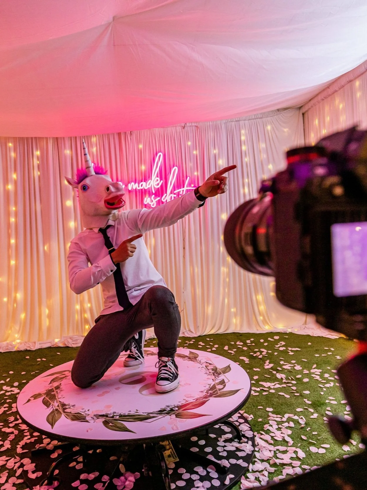 Person wearing unicorn mask posing indoors with camera focusing on them, surrounded by fairy lights and a pink neon sign, standing on decorated platform with scattered rose petals.