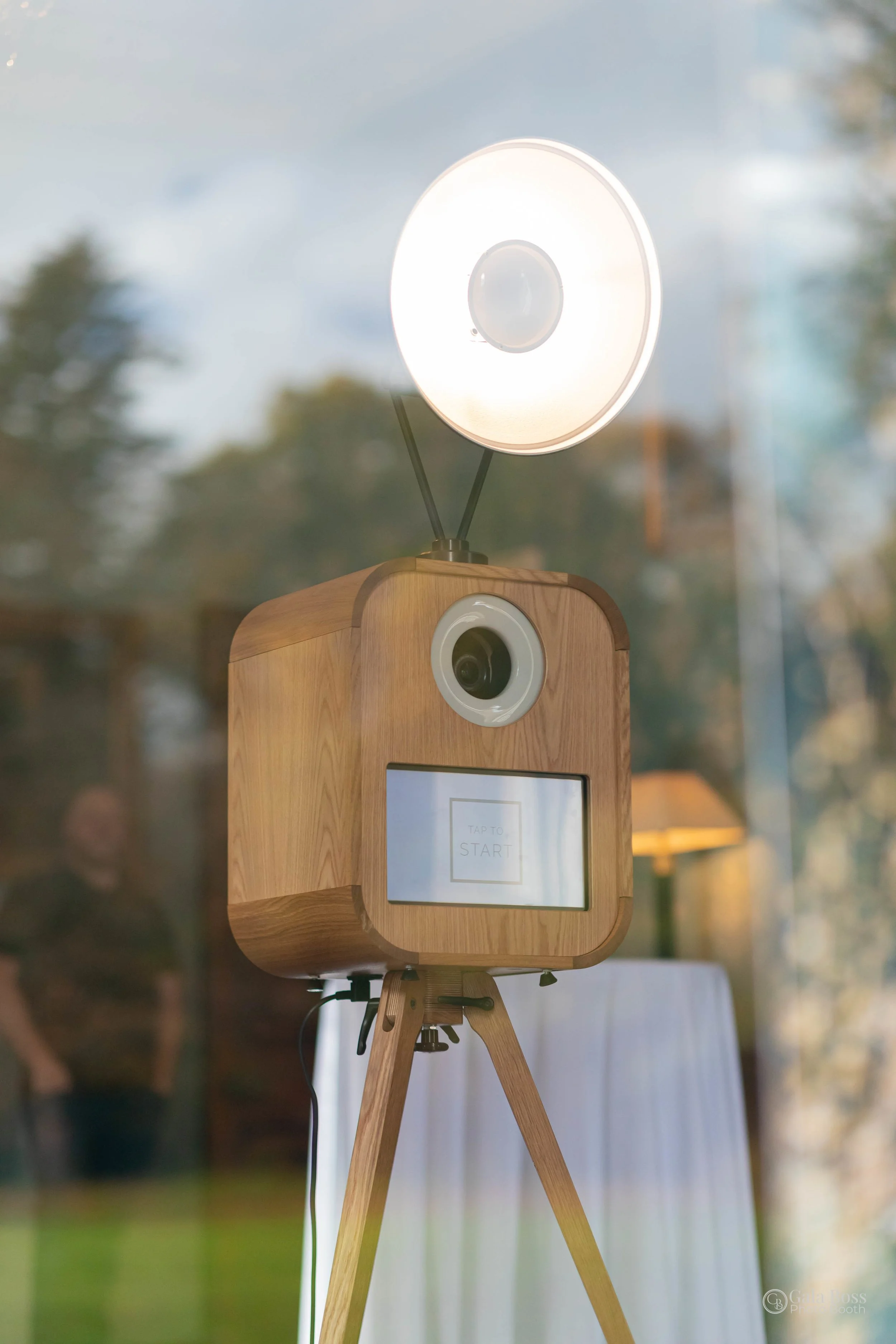 A wooden photo booth with a camera, a ring light, and a touchscreen, set up indoors near a window with reflections of trees outside.