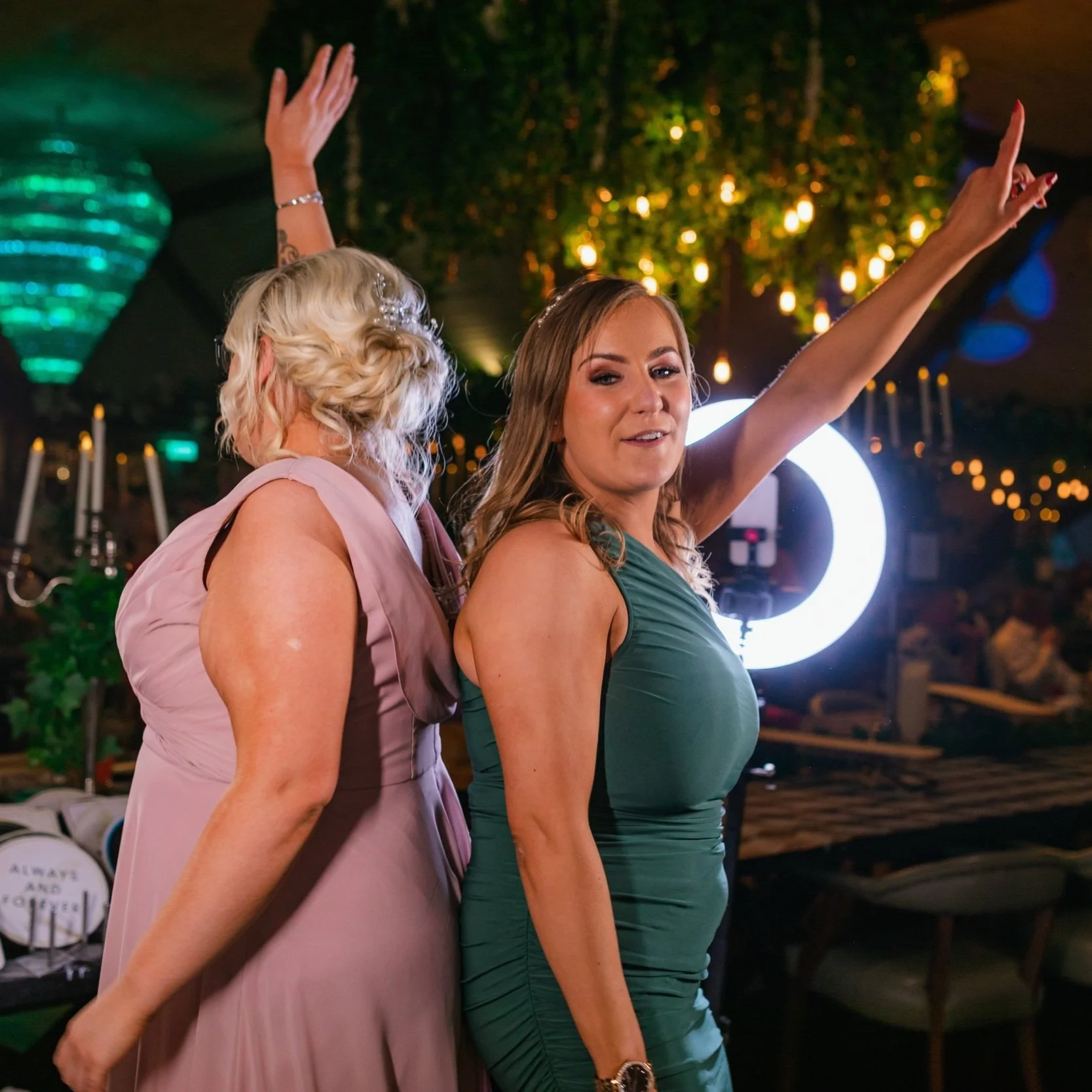 Two women dancing and enjoying a celebration in a decorated indoor venue with hanging lights and greenery.