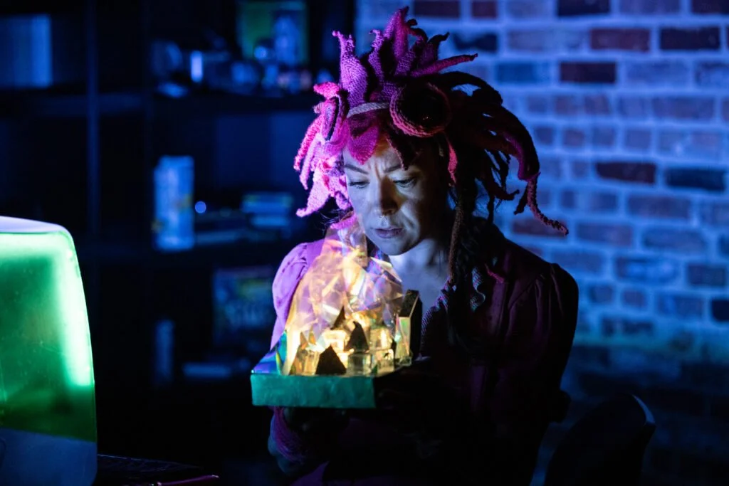 Woman with pink dreadlocks looking at a holographic display in a dimly lit room with a brick wall background.