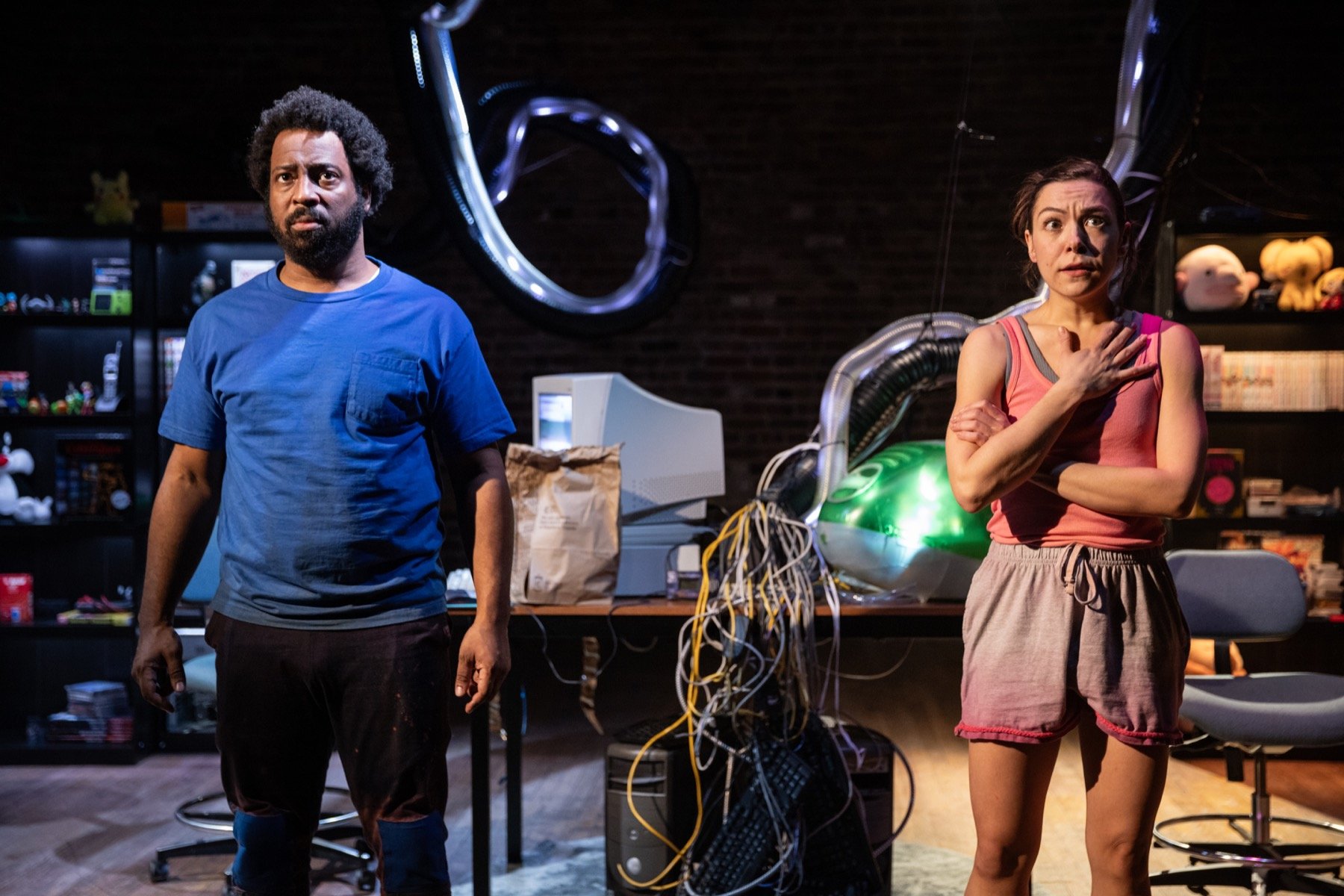 Two people, a man with a beard and a woman, stand in a cluttered room with wires, computers, and space-themed decorations, appearing surprised or concerned.