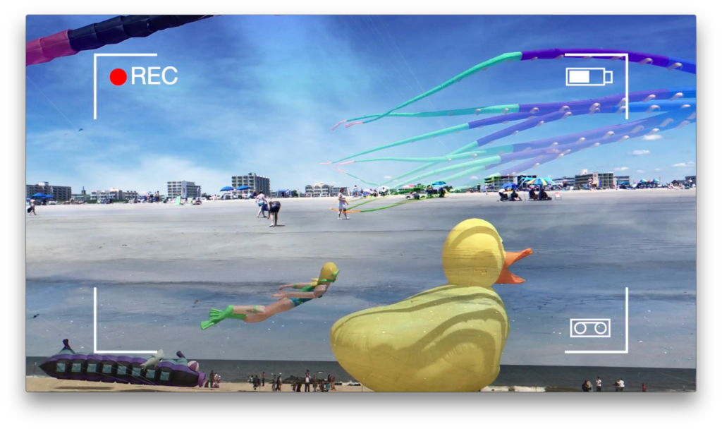 Photo of a beach scene with buildings in the distance, taken through a camera viewfinder overlay. Visible elements include a large inflatable rubber duck, a yellow and green inflatable rubber duck and a smaller yellow rubber duck, a woman with a yell