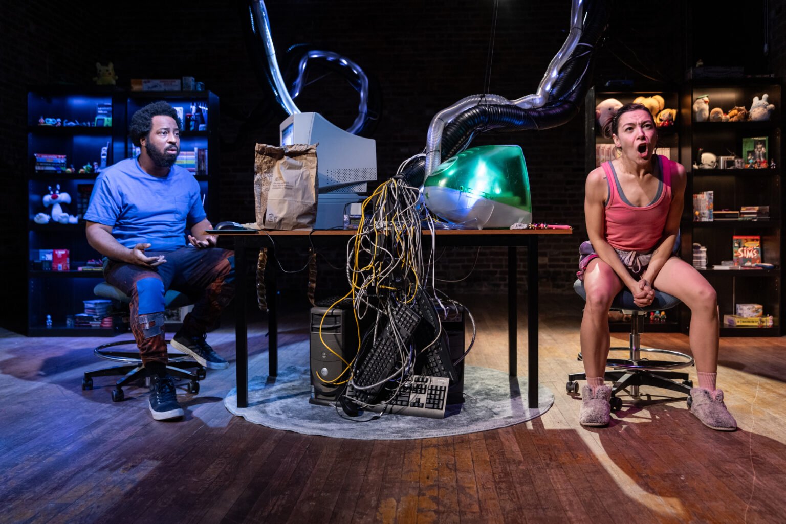 A man and a woman sit in an empty room with a chaotic table between them. The man on the left, with curly hair and a blue shirt, looks puzzled. The woman on the right, with dark hair and a pink top, appears angry or upset. The table has tangled wires