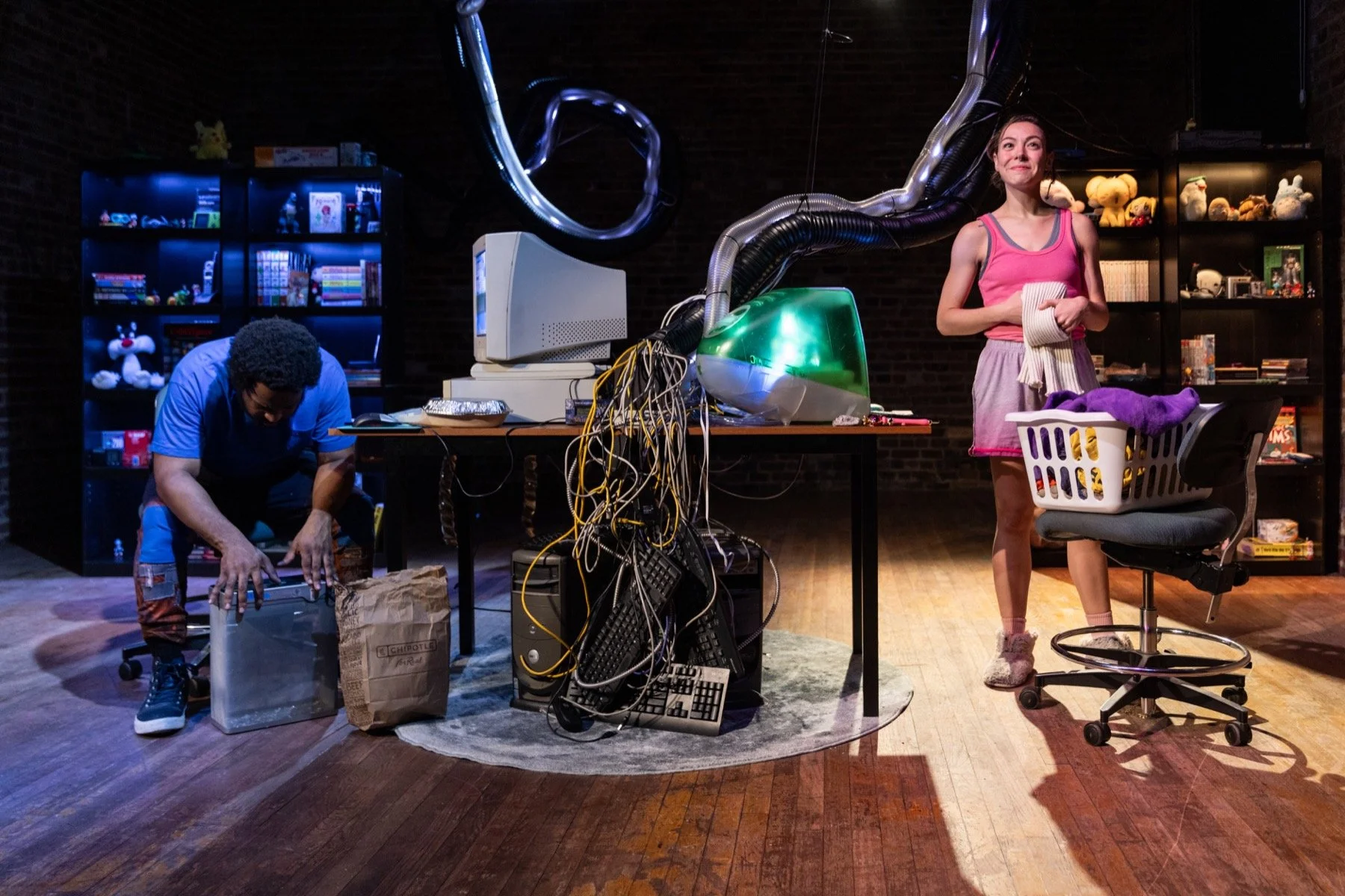 Two people organizing a cluttered workspace with electronic equipment and stuffed animals. The person on the left, wearing a blue shirt, is crouching and reaching into a paper bag, while the person on the right, wearing a pink tank top and shorts, is