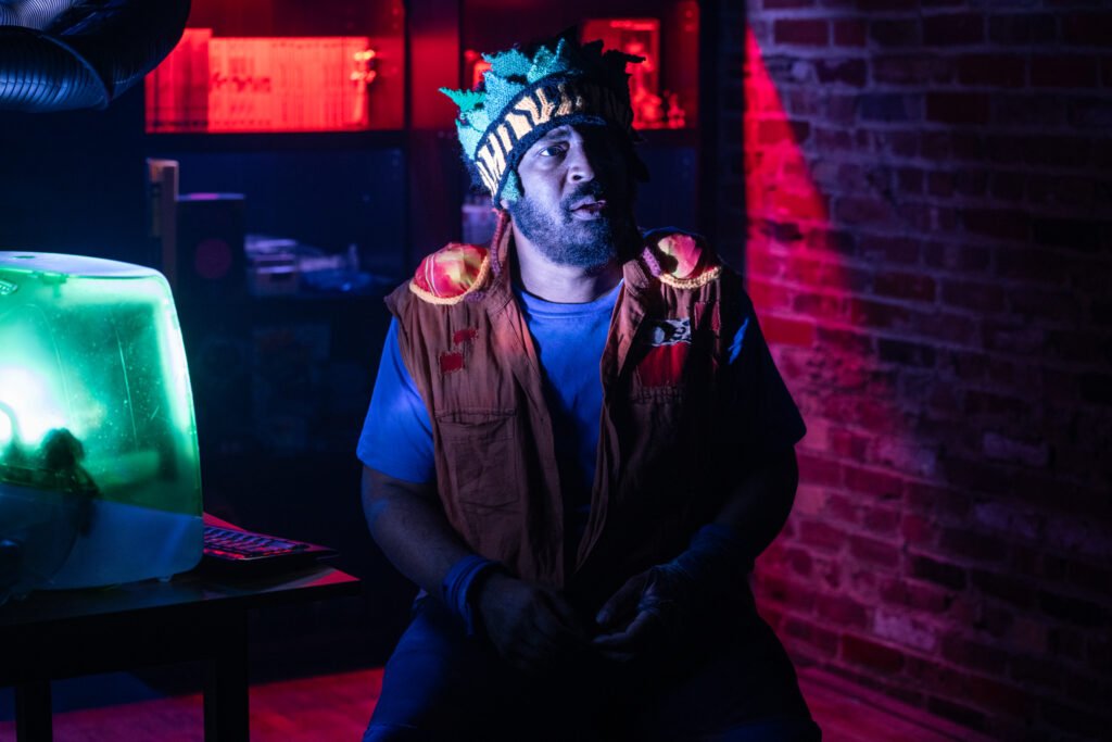 A man with a beard wearing a colorful mask and a brown vest sitting in a dimly lit room with neon red and green lighting, next to a computer monitor.