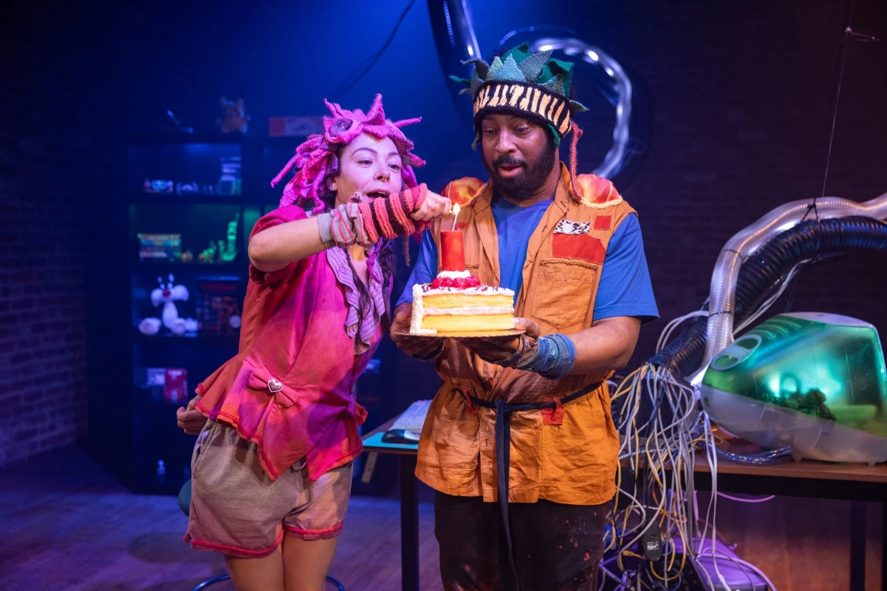 Two people celebrating with a birthday cake, one woman with pink dreadlocks and a colorful outfit and a man wearing a hat with the word 'Victory' on it, holding the cake with a lit candle.