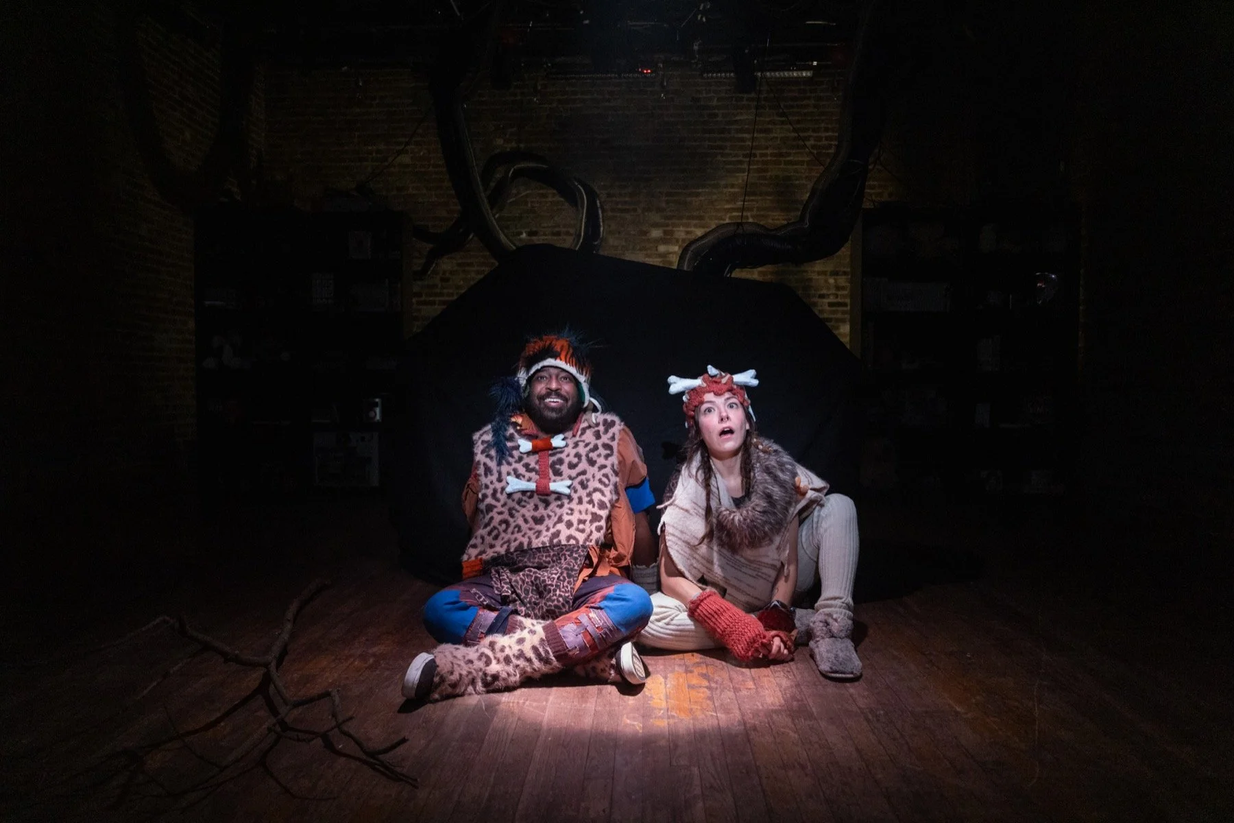 Two actors dressed in tribal costumes sit on the floor in a theatrical setting with dark lighting, brick walls, and a large black backdrop.